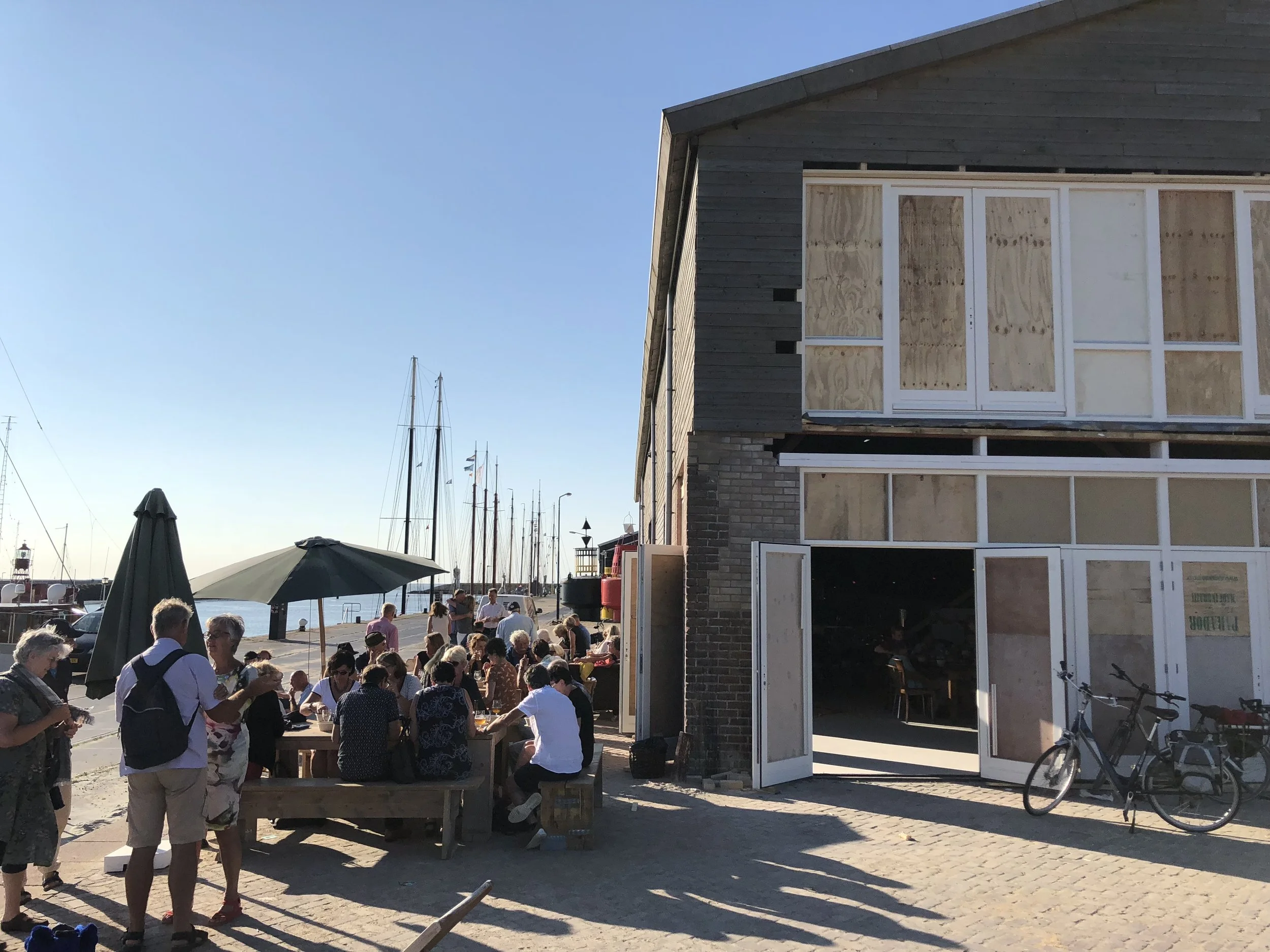 Een groep mensen die samen buiten aan tafels zitten en genieten van een zonnige dag bij een haven met schepen.