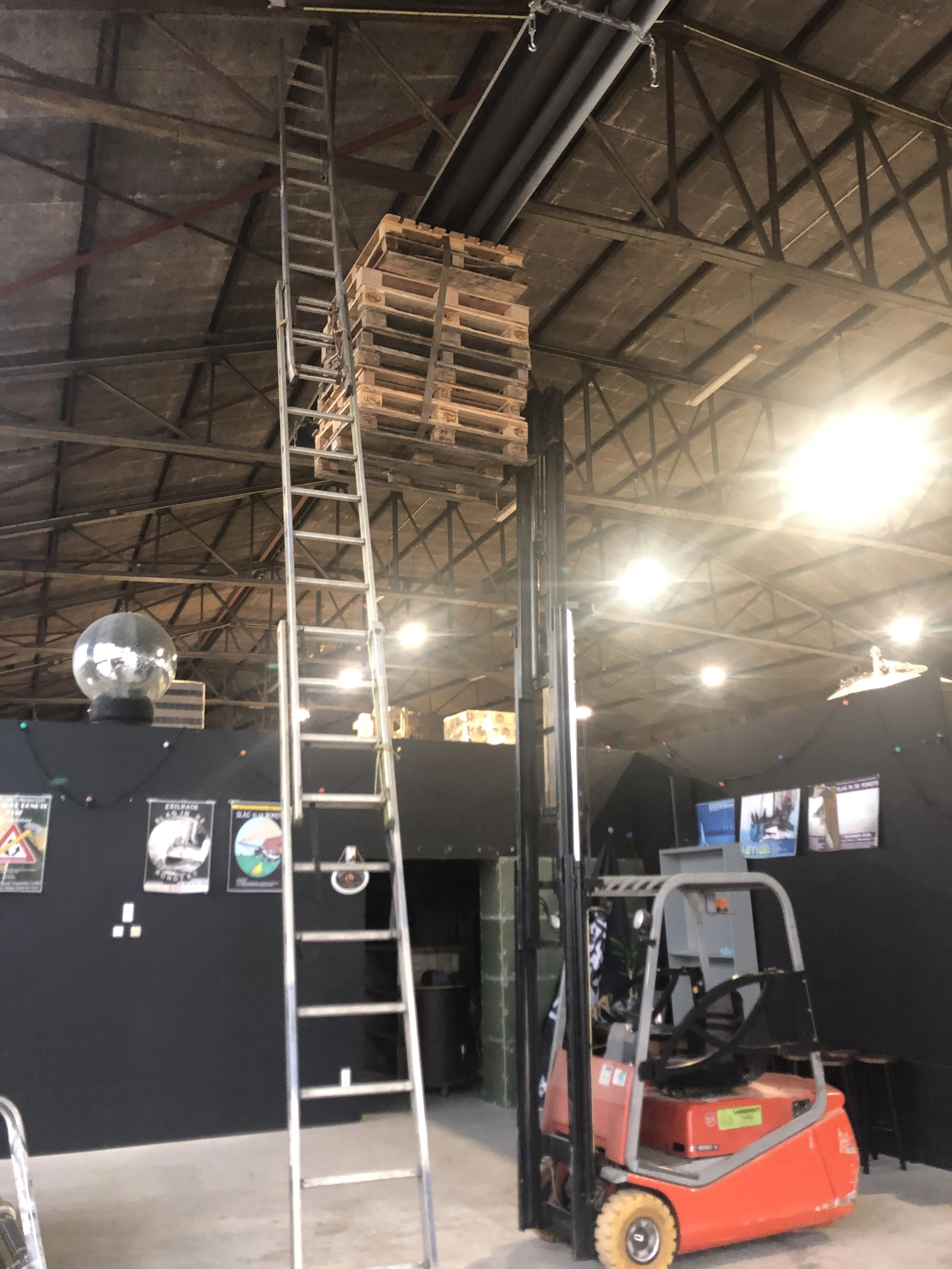 In een binnenruimte wordt een stalen ladder gebruikt om pallets op te tillen naar het plafond aan een ketting, terwijl een vorkheftruck zich onderaan bevindt.
