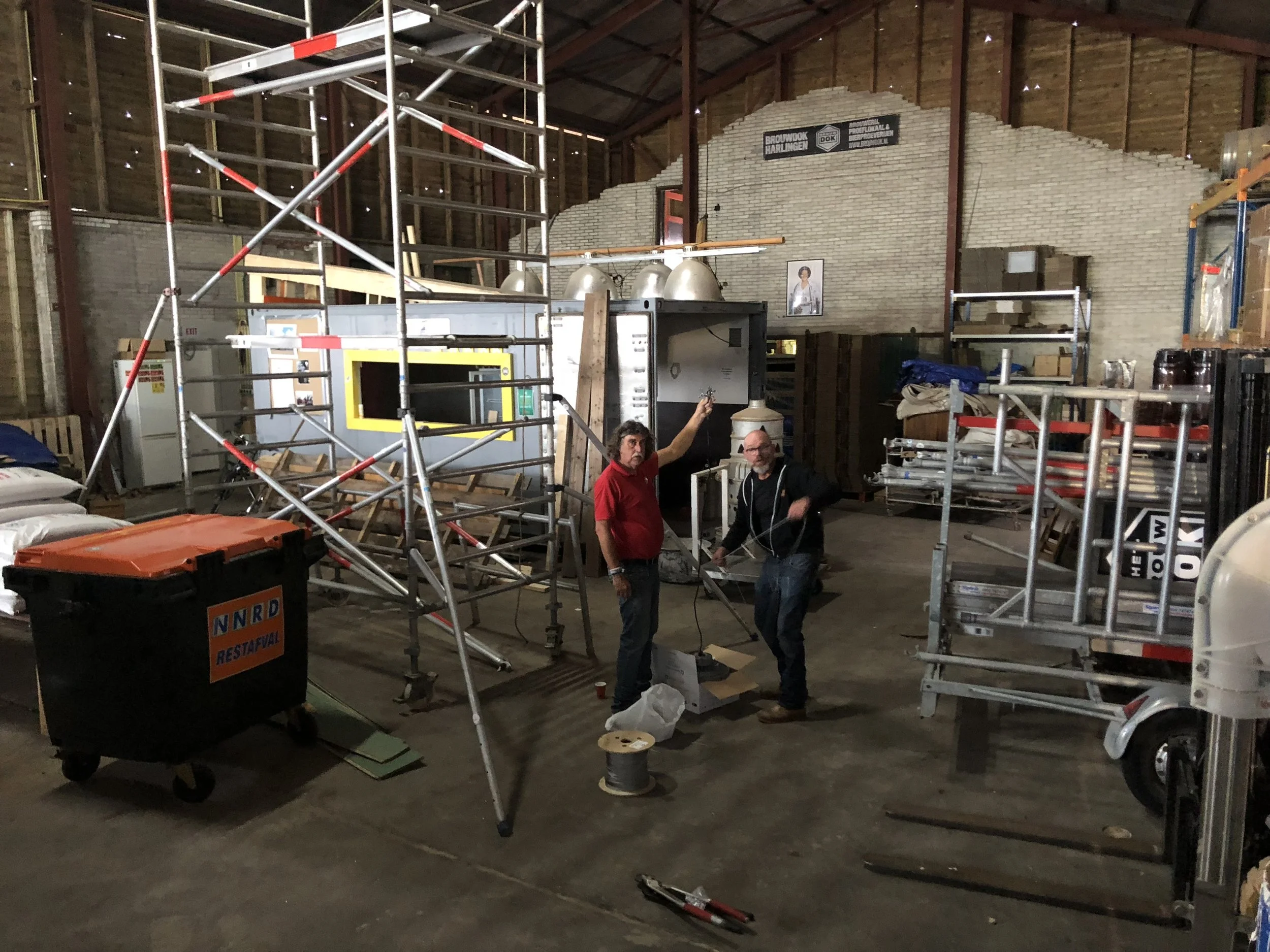 Binnen in een schuur met metalen rekken, werktafel met mensen die aan het werk zijn, deels gebouwd met bakstenen.