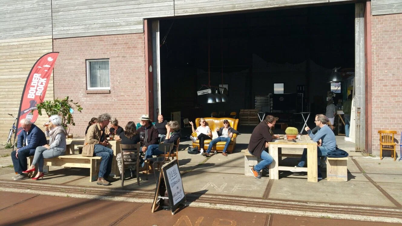 Mensen zitten buiten in een voorruimte onder een grote open deur, met een bord en een vlag met de tekst 'BOLDER ROCK', en er staan verschillende stoelen en tafels.