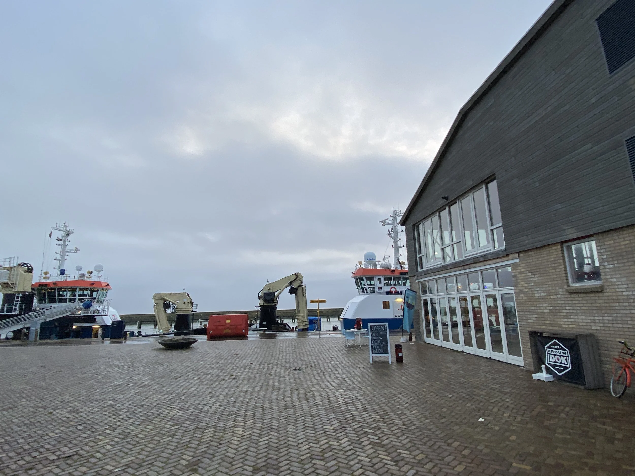 Een haven met boten en schepen, een modern gebouw met grote ramen, en een nog bewolkte hemel.