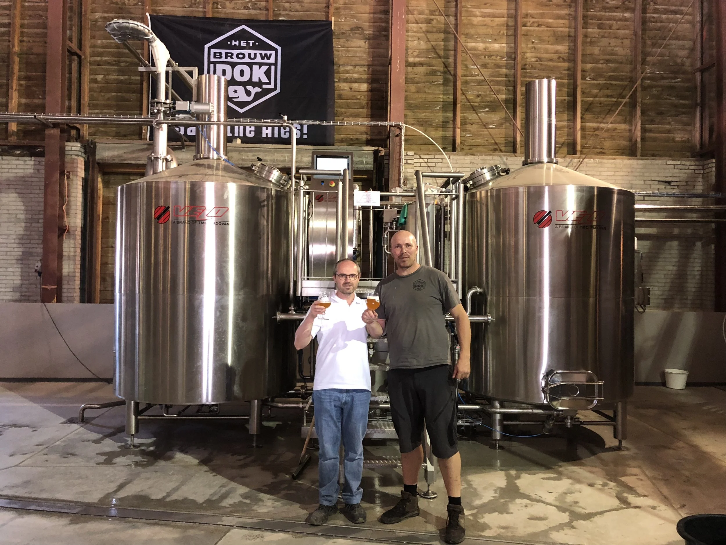Twee mensen staan voor grote bierketels in een brouwerij, beide houden een glas bier omhoog, een man draagt een wit handdoek en een ander een grijs shirt. Achter hen hangt een zwarte vlag met witte tekst en een logo.