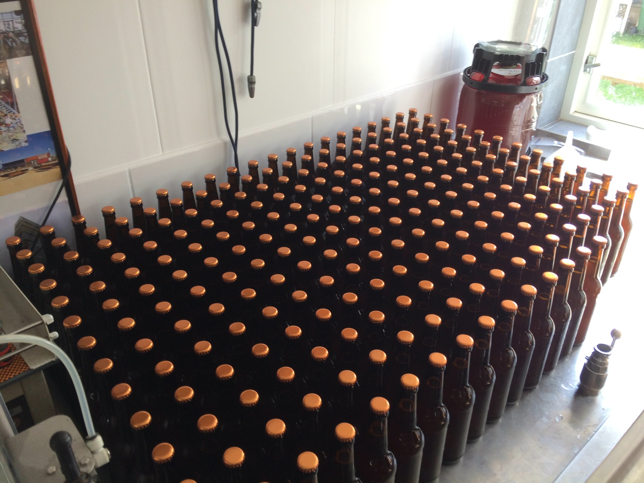 Rijen lege heldere bierflesjes met oranje doppen, op een tafel in een kamer met een witte muur, een grote ton en een deur naar buiten.
