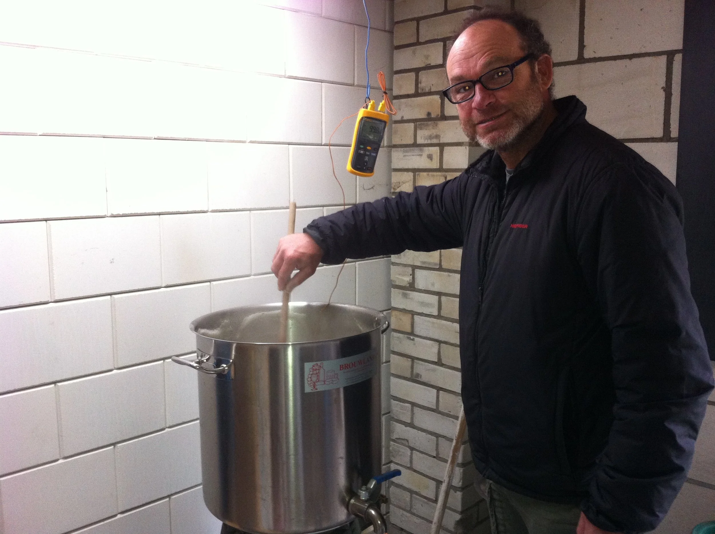 Man met bruin haar, een baard en bril, die bezig is met een witte vloeistof in een grote metalen container te gieten. De container heeft een logo en zit onder een elektrische meter met draden die eraan hangen, in een ruimte met witte tegels en een ba
