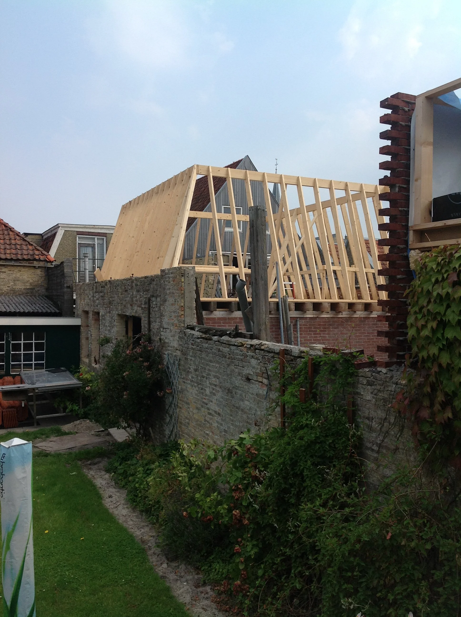 Een huis in aanbouw met een houten constructie voor het dak, omgeven door een tuin met groene planten en oude muren.