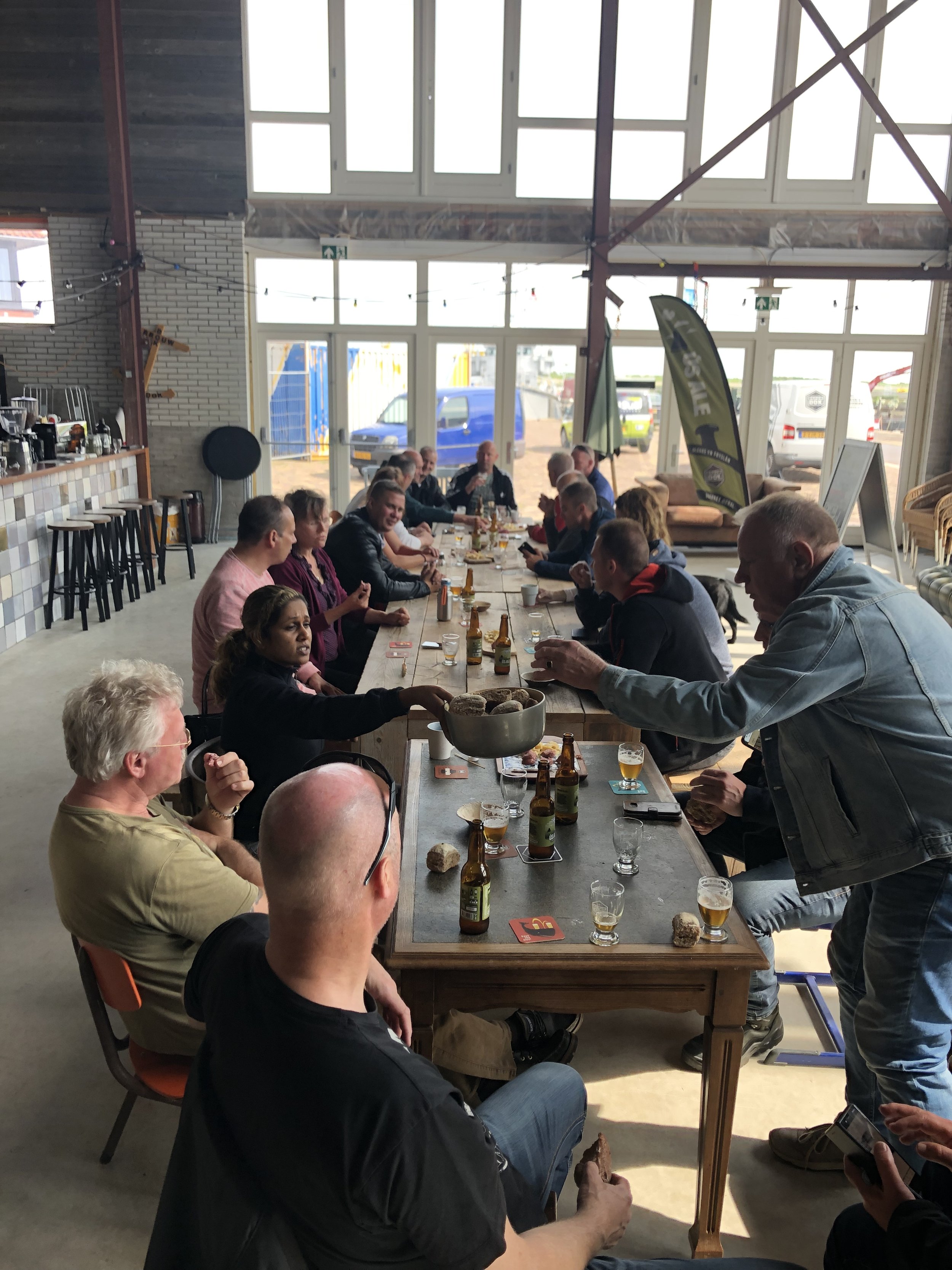 Een groep mensen zit aan een lange tafel in een lichte eetzaal, drinken en praten. Ze lijken deel uit te maken van een sociale bijeenkomst of bedrijfsfeest.