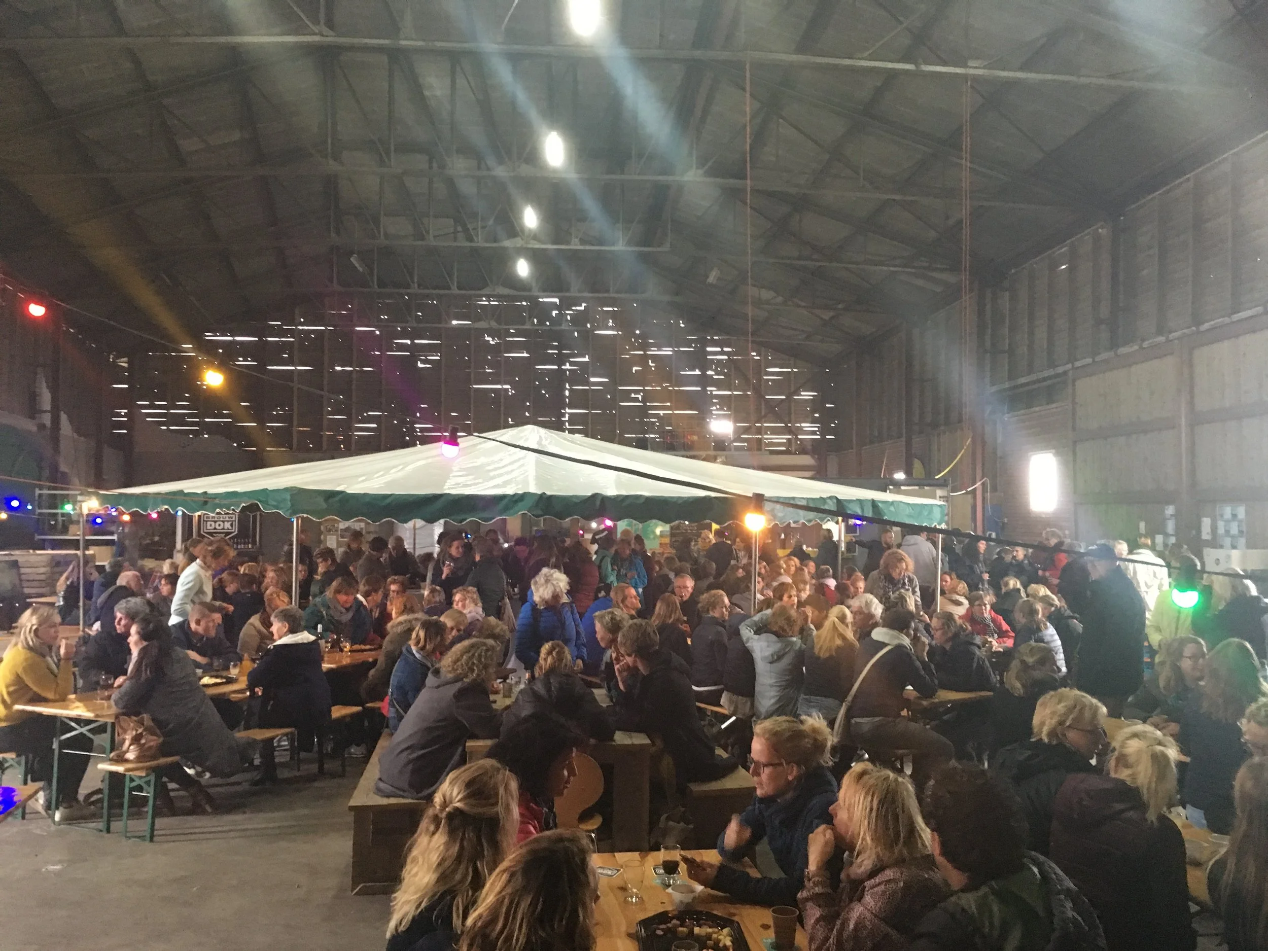Een drukke binnenruimte tijdens een evenement met mensen die aan lange tafels zitten, gedronken en praten. In de achtergrond een grote boog met licht en een muur met horizontale lichtgaatjes.
