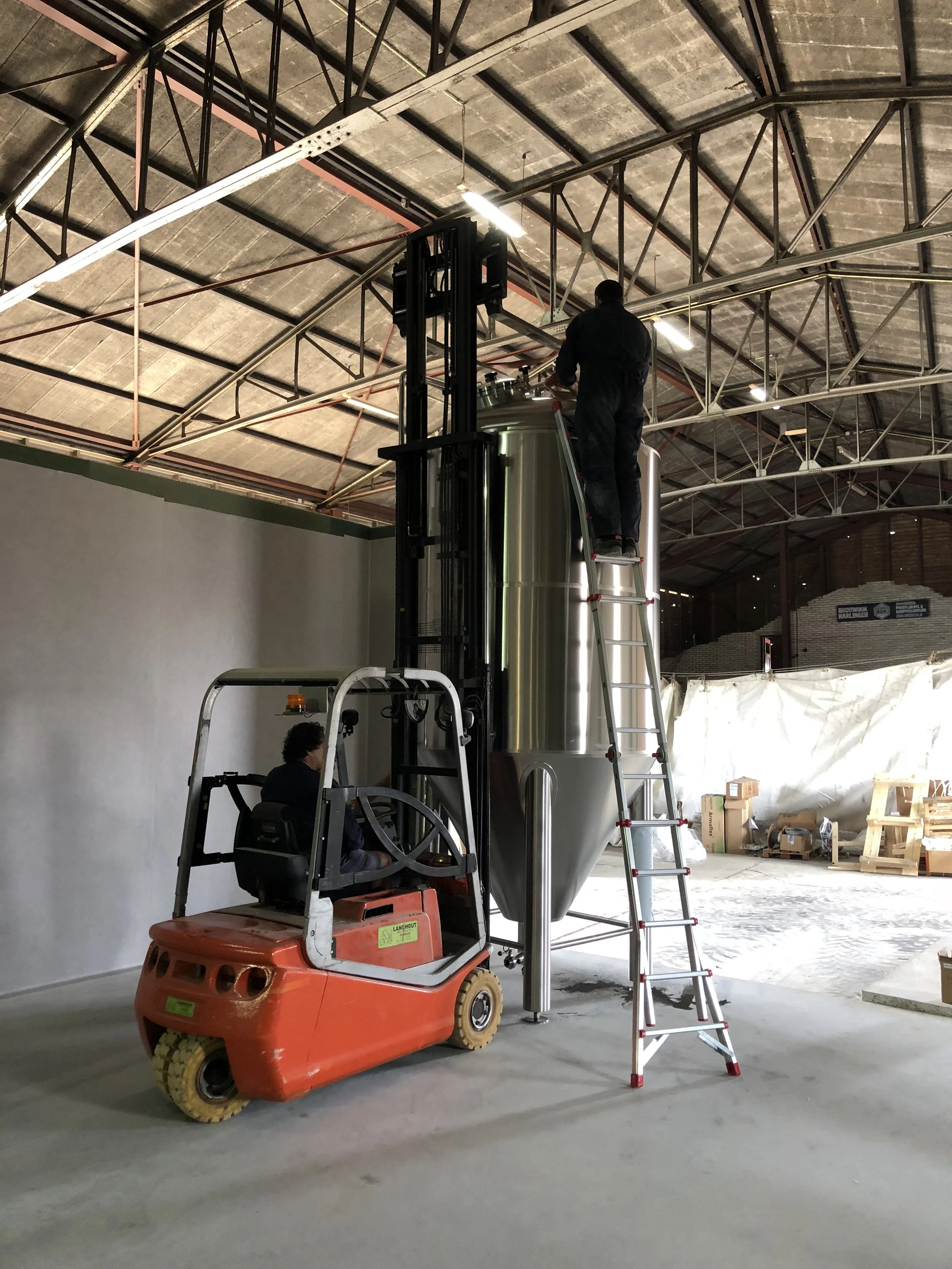 Een persoon werkt op een grote metalen tank met behulp van een heftruck en een steiger in een industrieel gebouw.