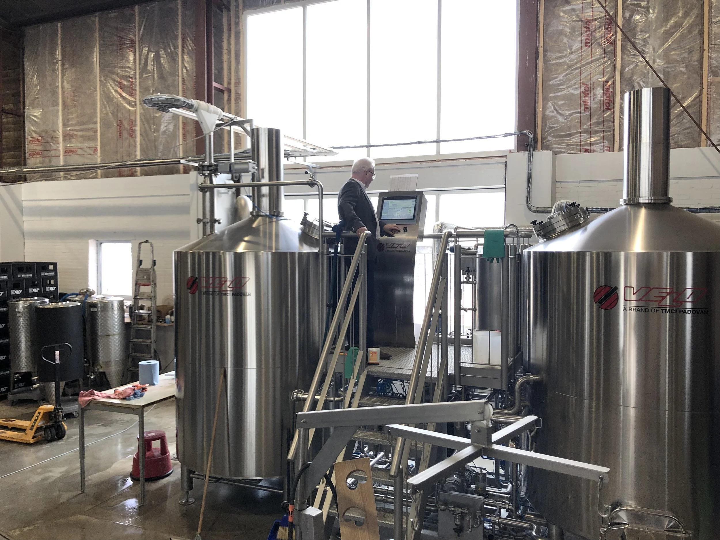 Binnen een industrieel gebouw met grote ramen, staat een man in pak bij palmatische industriële apparatuur.