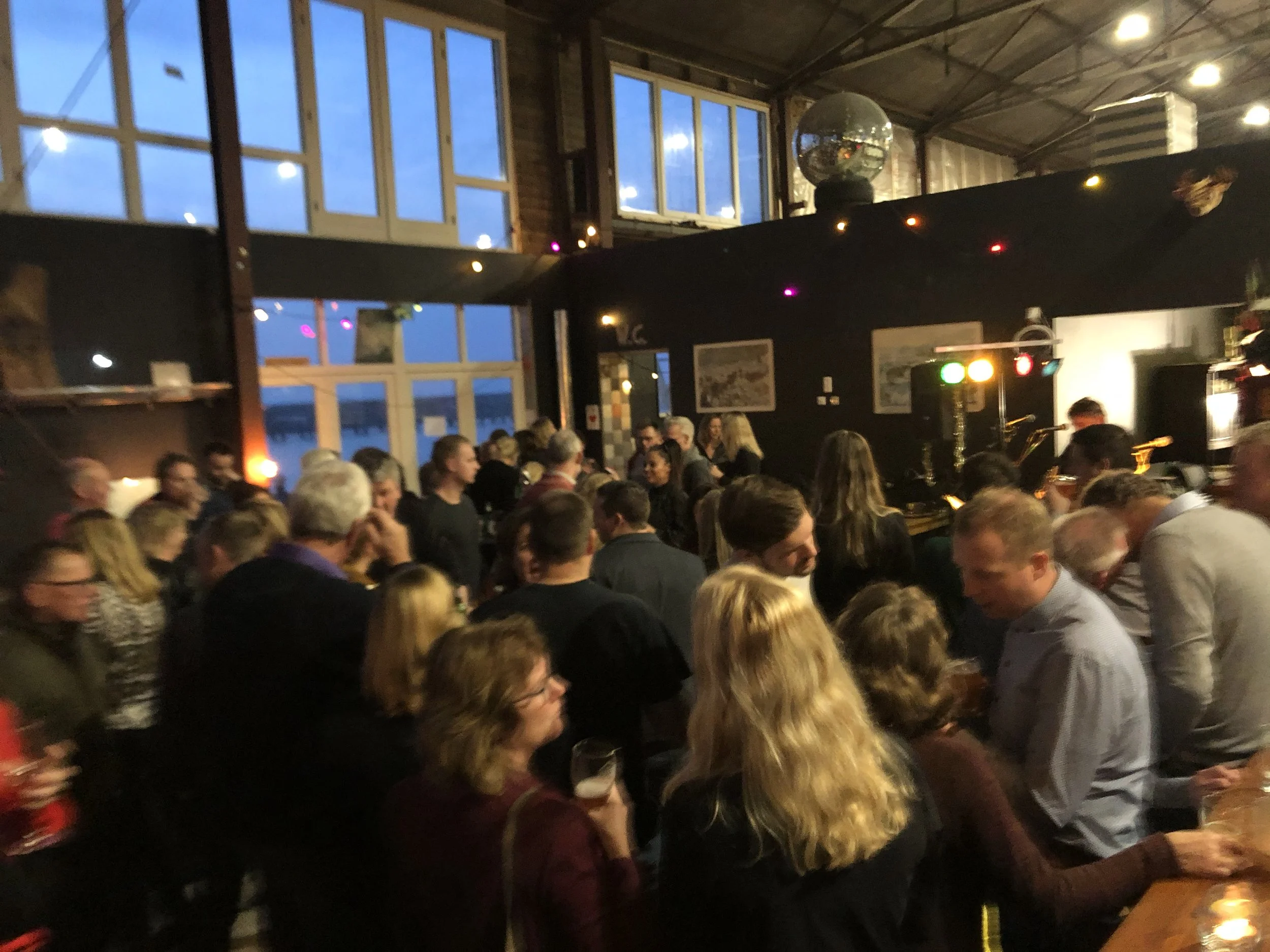 Binnen van een feestzaal met mensen die samen praten, op de achtergrond grote ramen en een discobal, lichteffecten en decoraties.