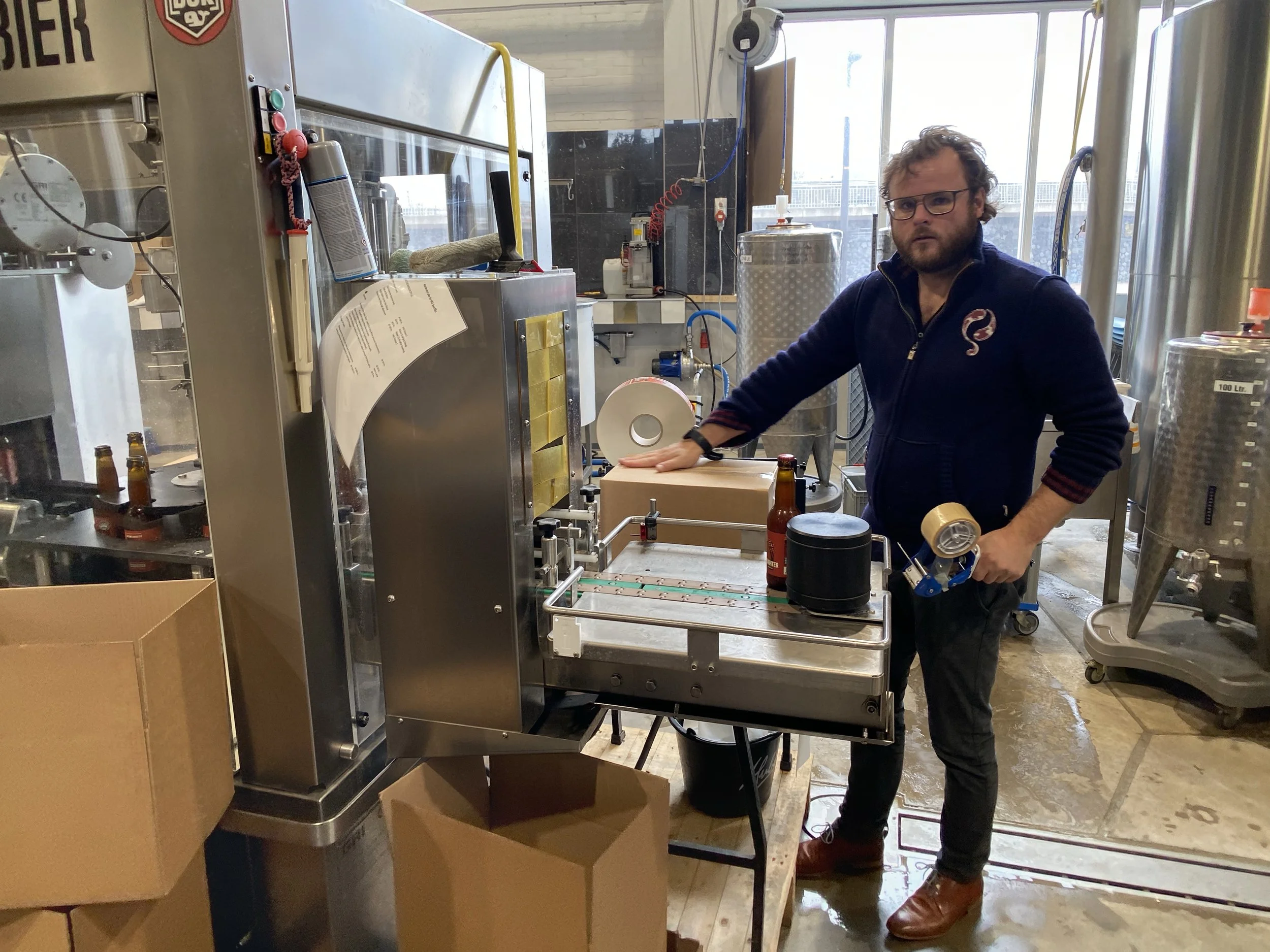 Man met een baard, bril en een donkerblauwe jas, staand bij een industriële machine in een fabriek, omringd door containers en gereedschap.