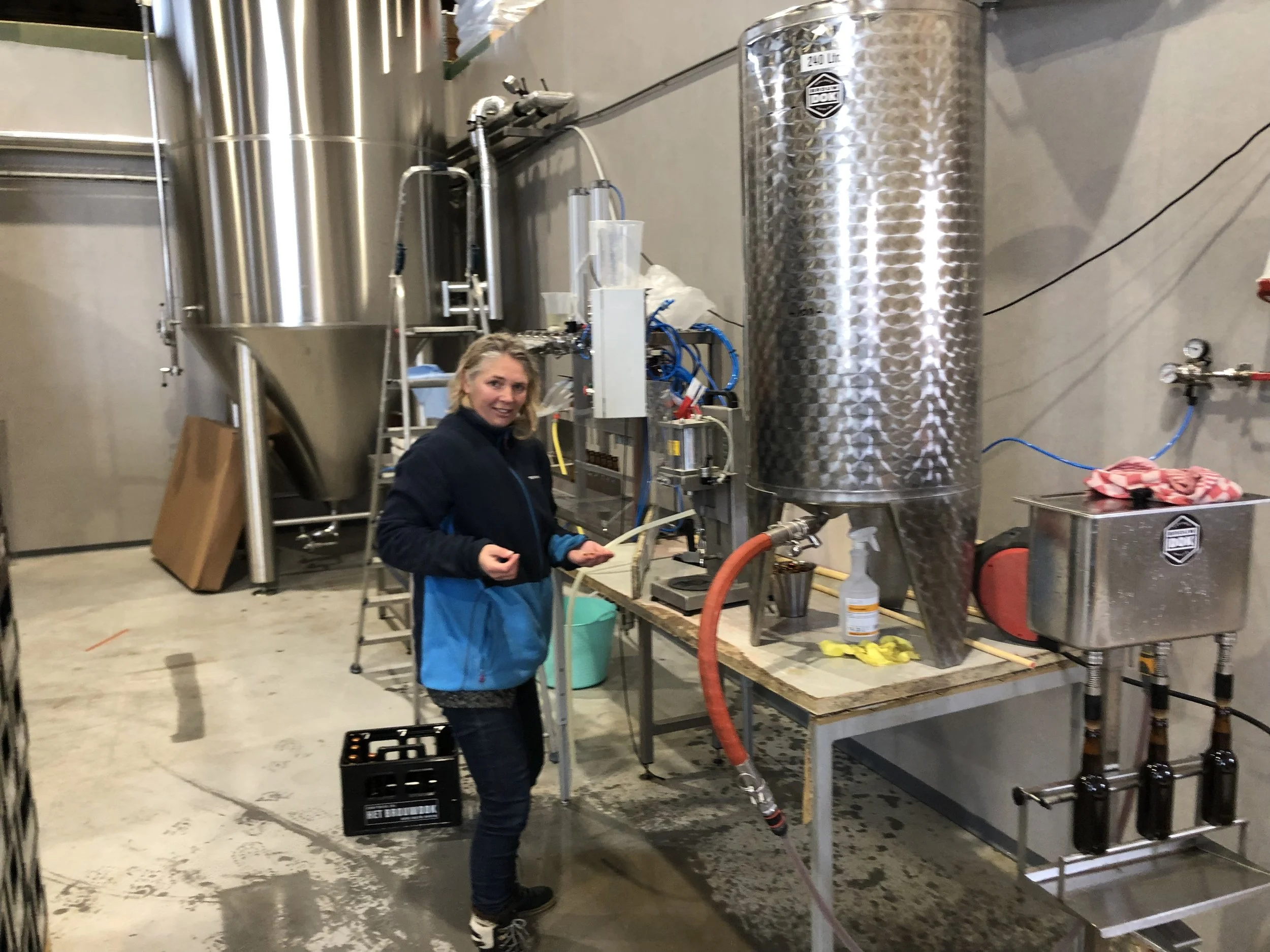 Vrouw in een industrieel laboratorium of brouwerij met grote metalen vaten en apparatuur, lachend naar de camera.