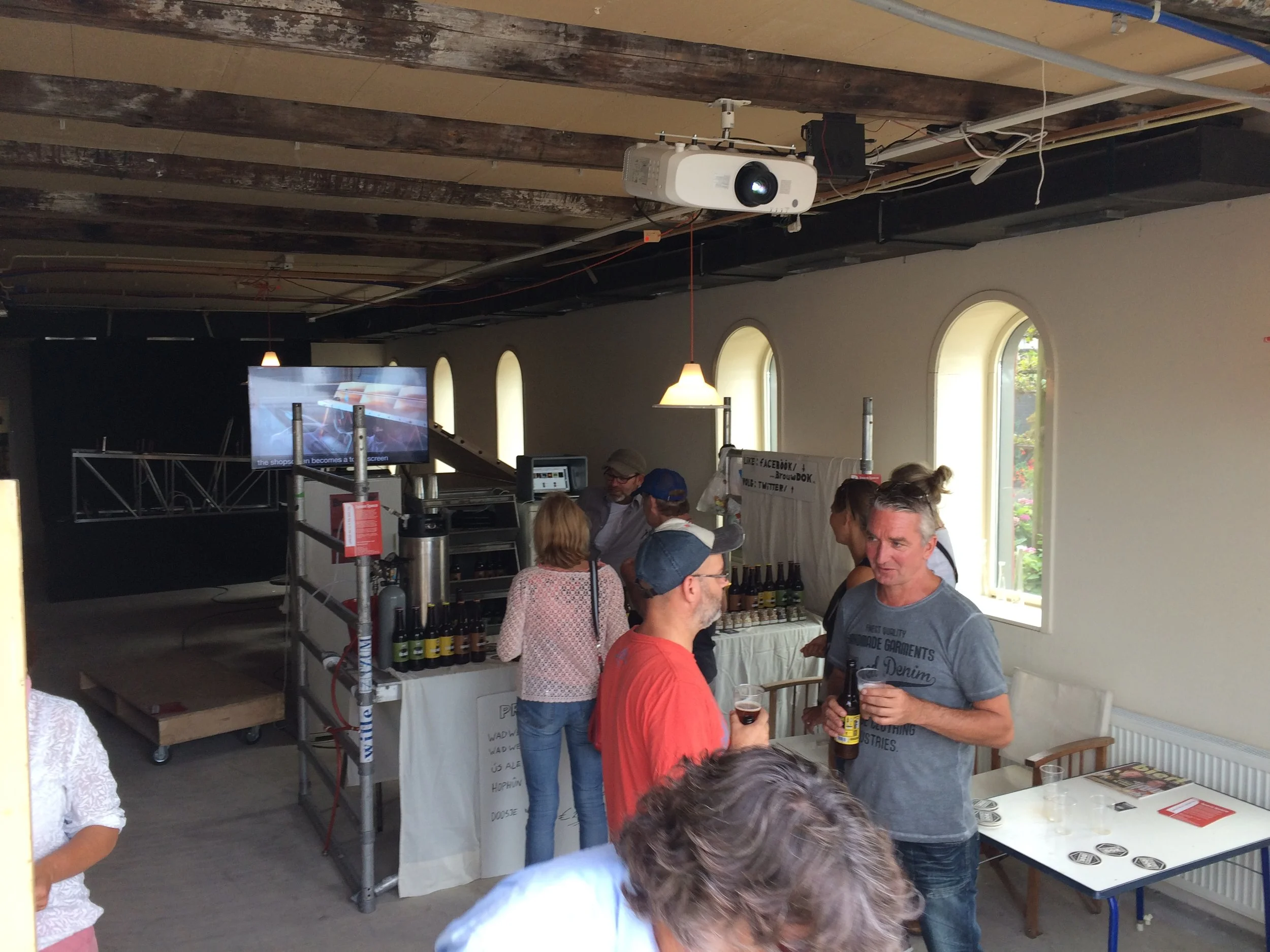 Binnenbeeld van een groep mensen die borrelen en praten bij een informele bijeenkomst, met een projector en een scherm op de achtergrond.