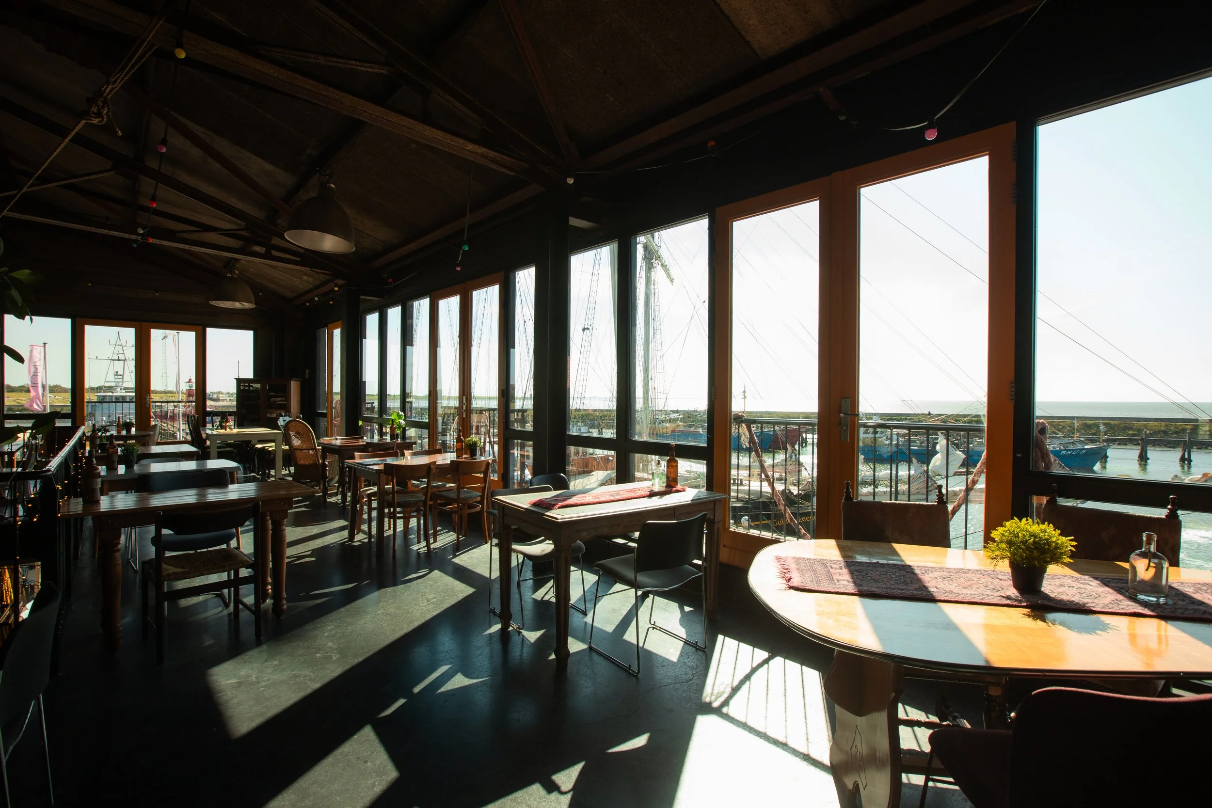 Interieur van een restaurant met grote ramen die uitkijken op een haven met boten, tafels en stoelen binnen.