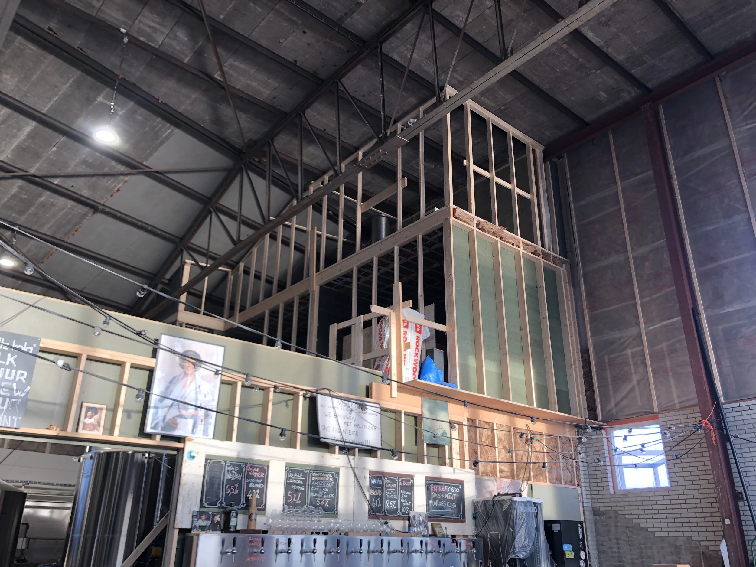 Binnenkant van een winkel of café onder constructie, met houten en metalen raamwerken, posters en menuborden, en een hoog plafond met geïnstalleerde verlichtingsarmaturen.