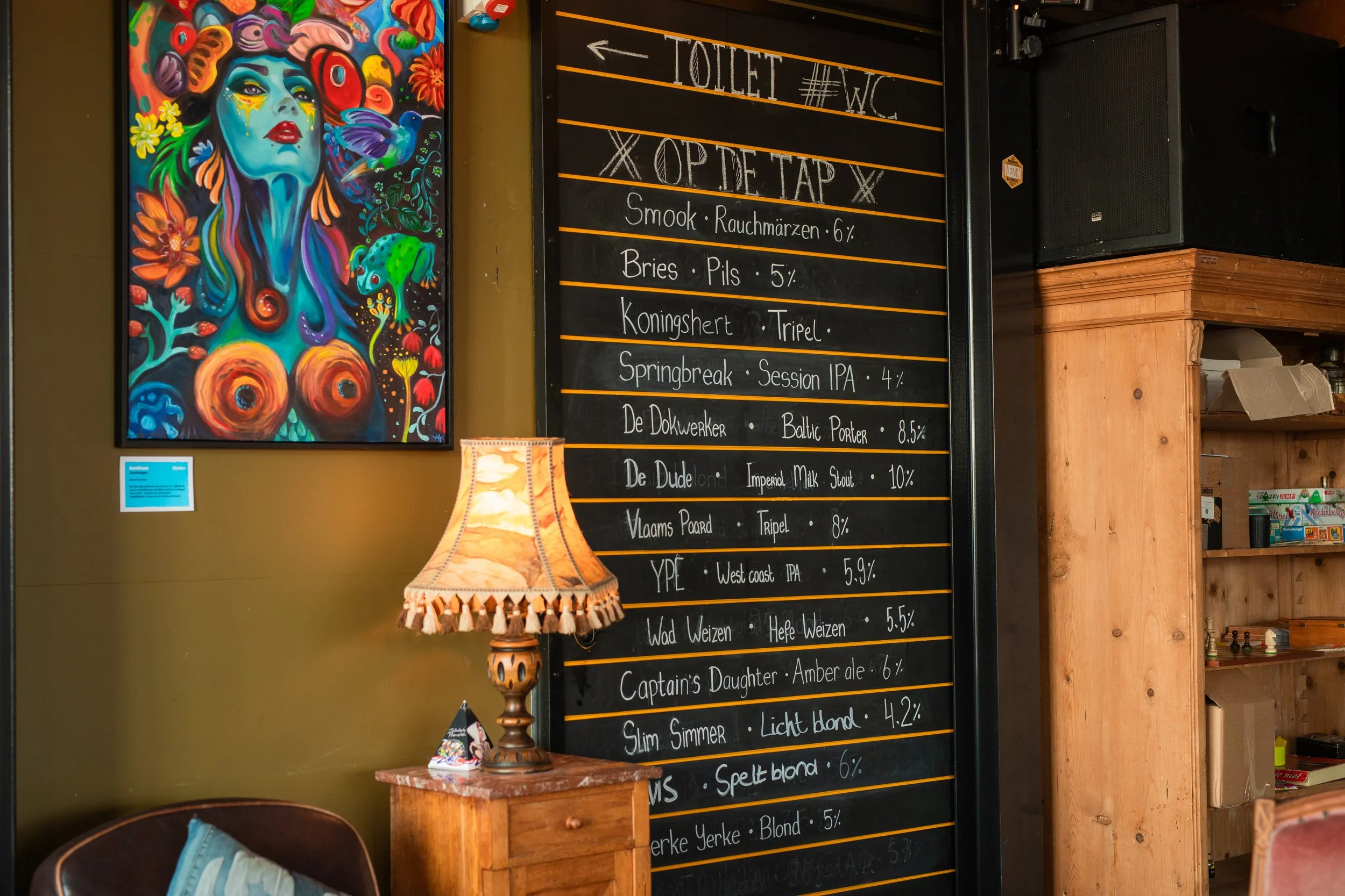 Een kleurrijk kunstwerk met een vrouwelijk gezicht en bloemen, naast een menu bord met drankprijzen in een café.