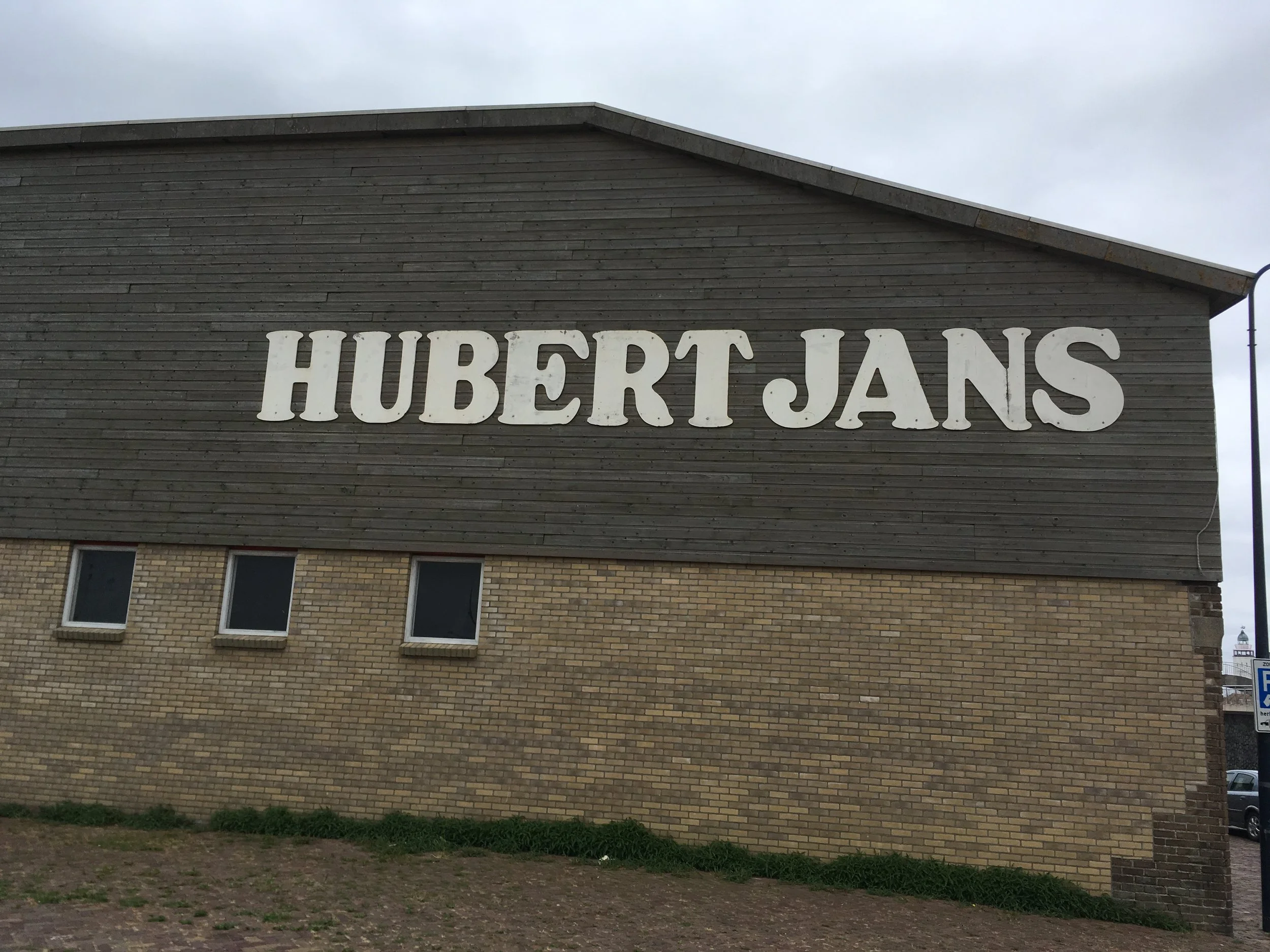Een gebouw met een houten bovenverdieping en een bakstenen onderverdieping, met grote witte letters die HUBERT JANS spellen.