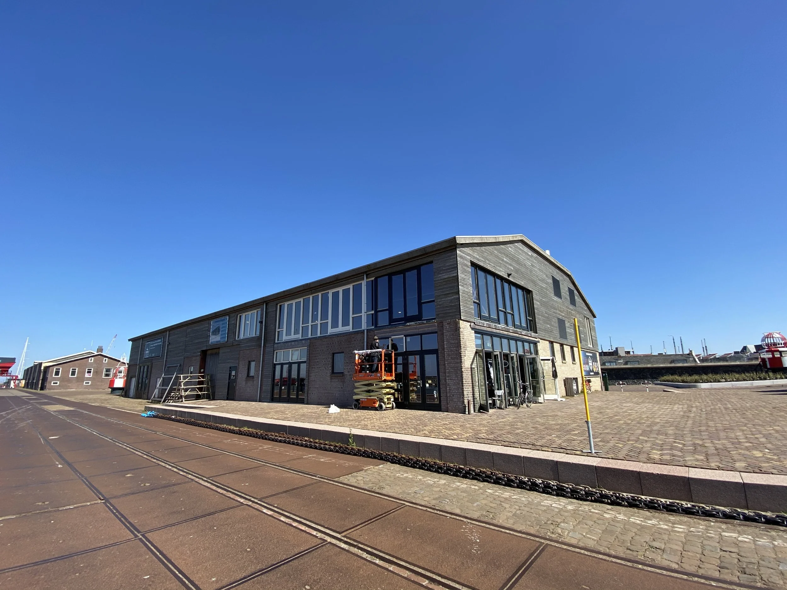 Nieuw gebouw onder constructie met een kraan, omringd door een parkeerterrein en straat, met een heldere blauwe lucht erboven.