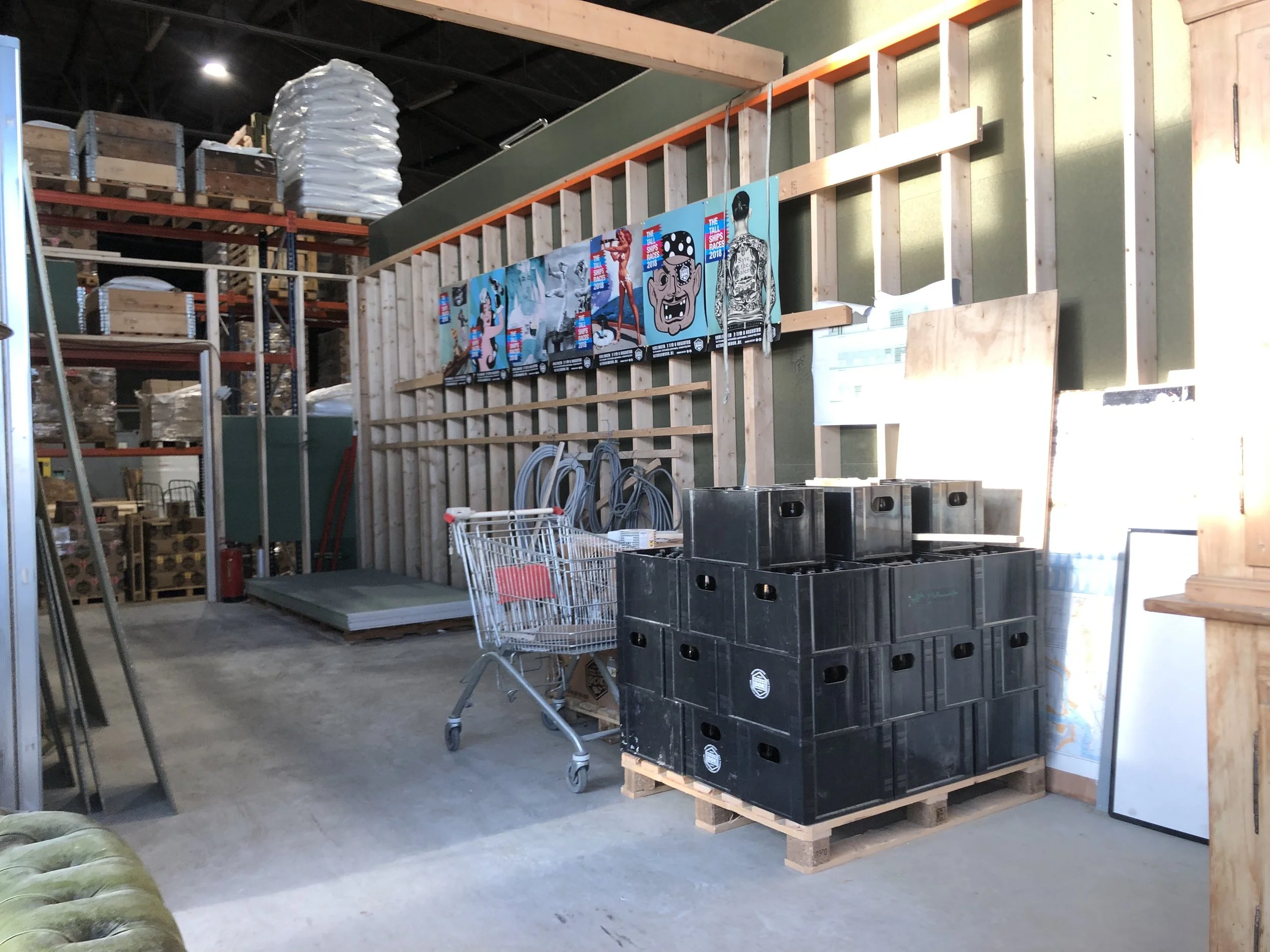 Interieur van een magazijn met houten stellingen, plastic bakken op pallets, een winkelwagen, posters aan de muur, en een lege ruimte met cementvloer.