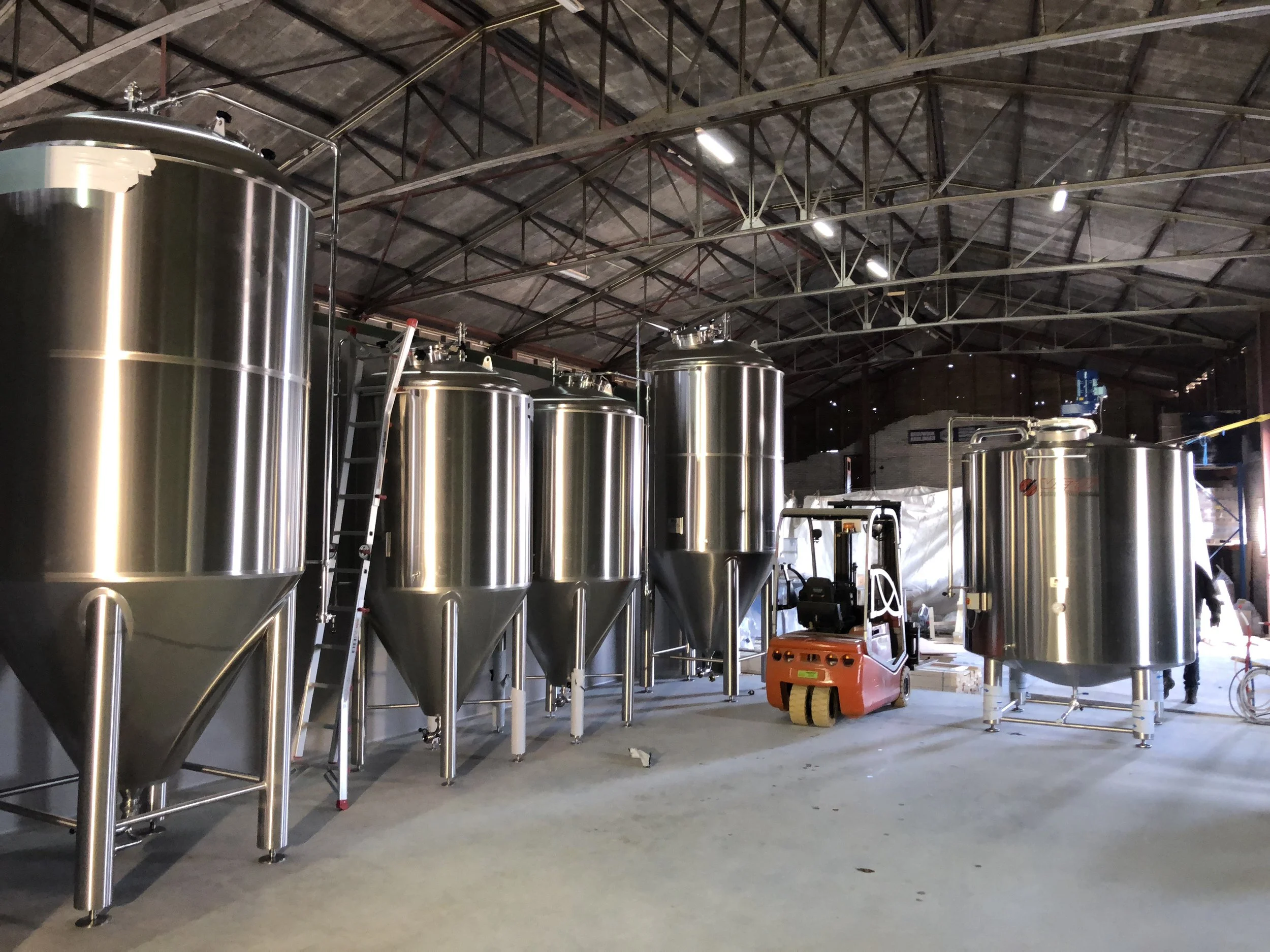 In een industrieel gebouw staan grote metalen tanks en apparatuur, waarschijnlijk voor het brouwen van bier of een soort productieproces. Er is een heftruck en bouwmaterialen zichtbaar, wat aangeeft dat de locatie nog in aanbouw of renovatie is.