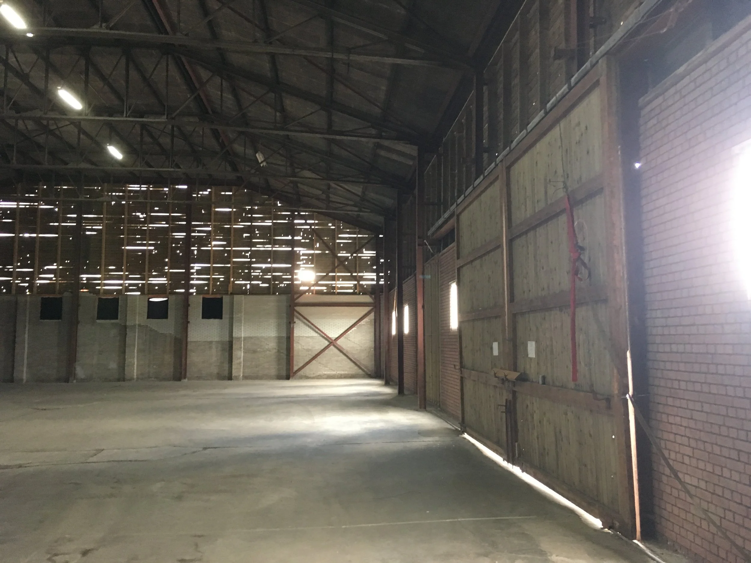 Binnenkant van een grote, lege loods of warehouse met een open structuur, houten en metalen muren en een betonnen vloer, met licht dat door kieren in het dak schijnt.