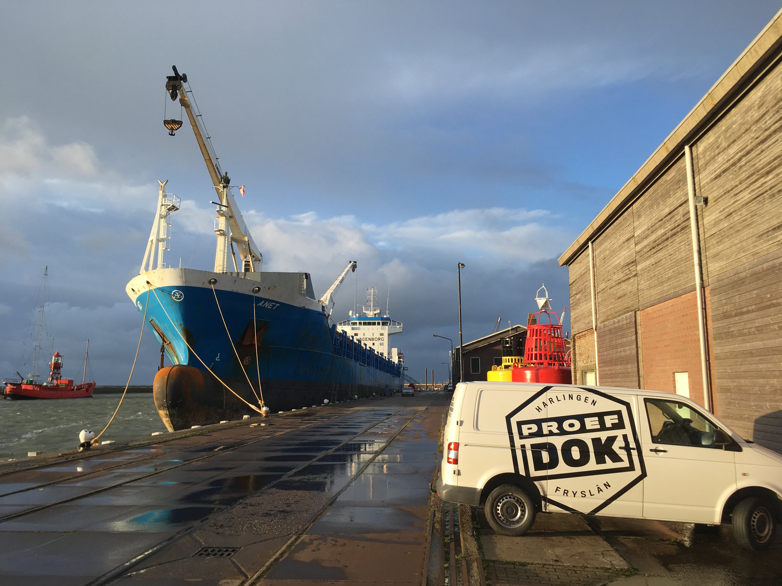 Een haven met schepen en boten, waaronder een grote blauwe schip met een hijskraan, en een witte bus met opschrift 'PROEF DOK' uit Friesland. Het weer is bewolkt en het lijkt te regenachtig.
