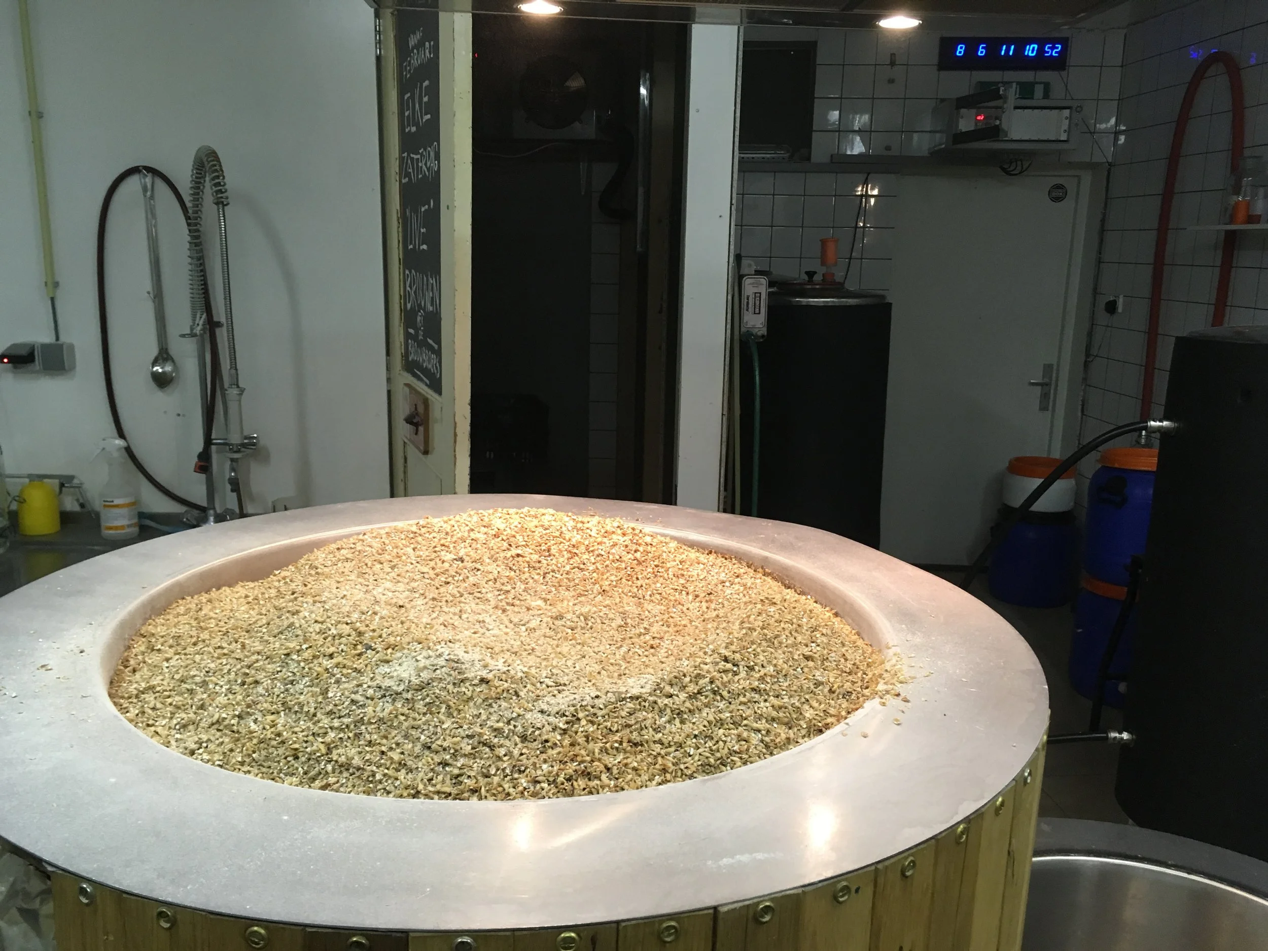 Ronde tafel met een mengsel van zaden of granen in een bak, in een keuken met witte tegels en verschillende apparatuur.