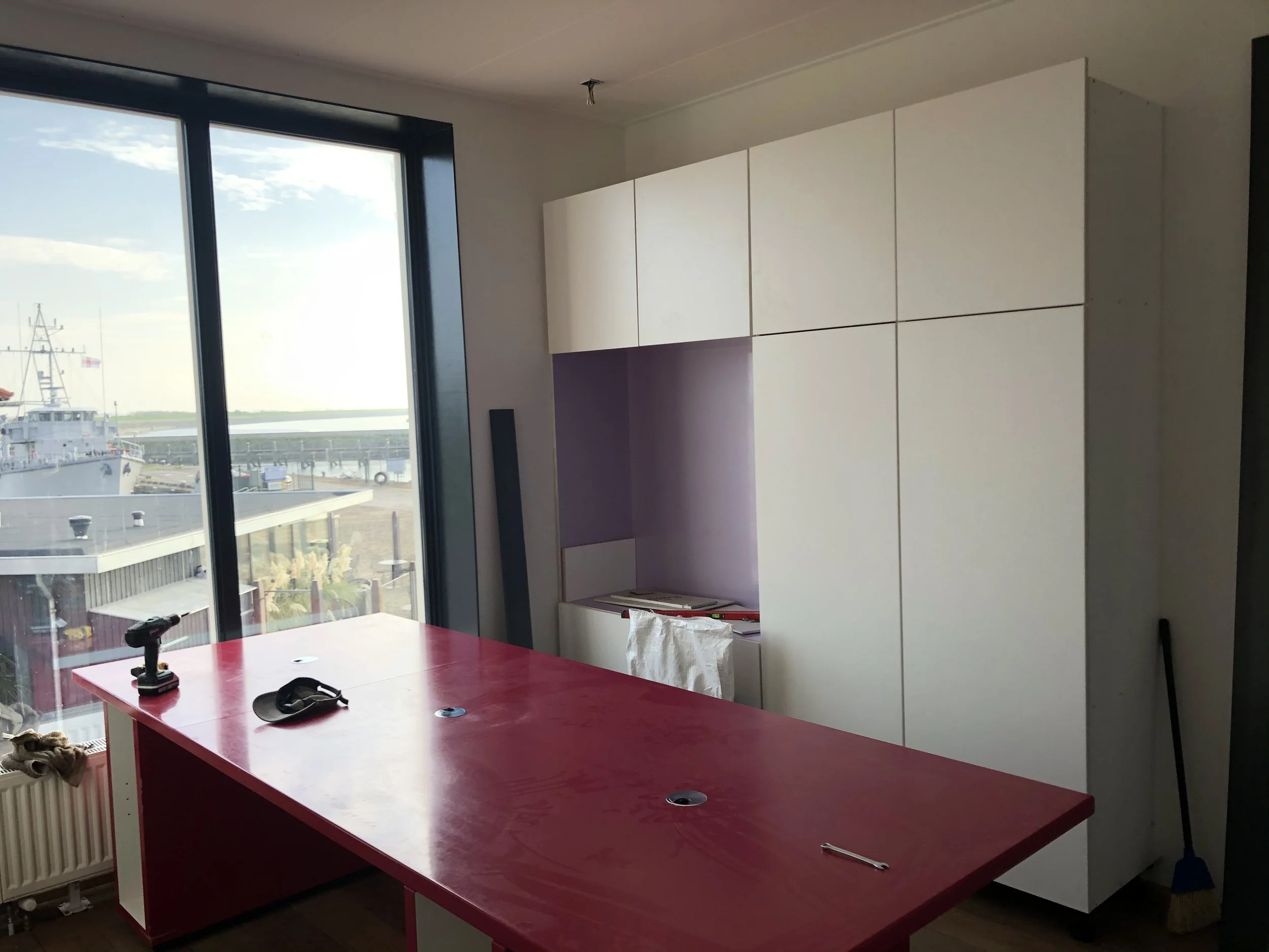 Binnenzicht van een woning met grote glazen ramen die uitkijken op een haven en een boot. Een rode tafel met enkele gereedschappen en een witte kast tegen de muur.