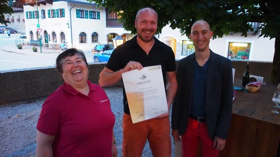 Drie mensen lachen en poseren met een certificaat, twee mannen en een vrouw, buiten onder een boom, op een terras met een stad op de achtergrond.