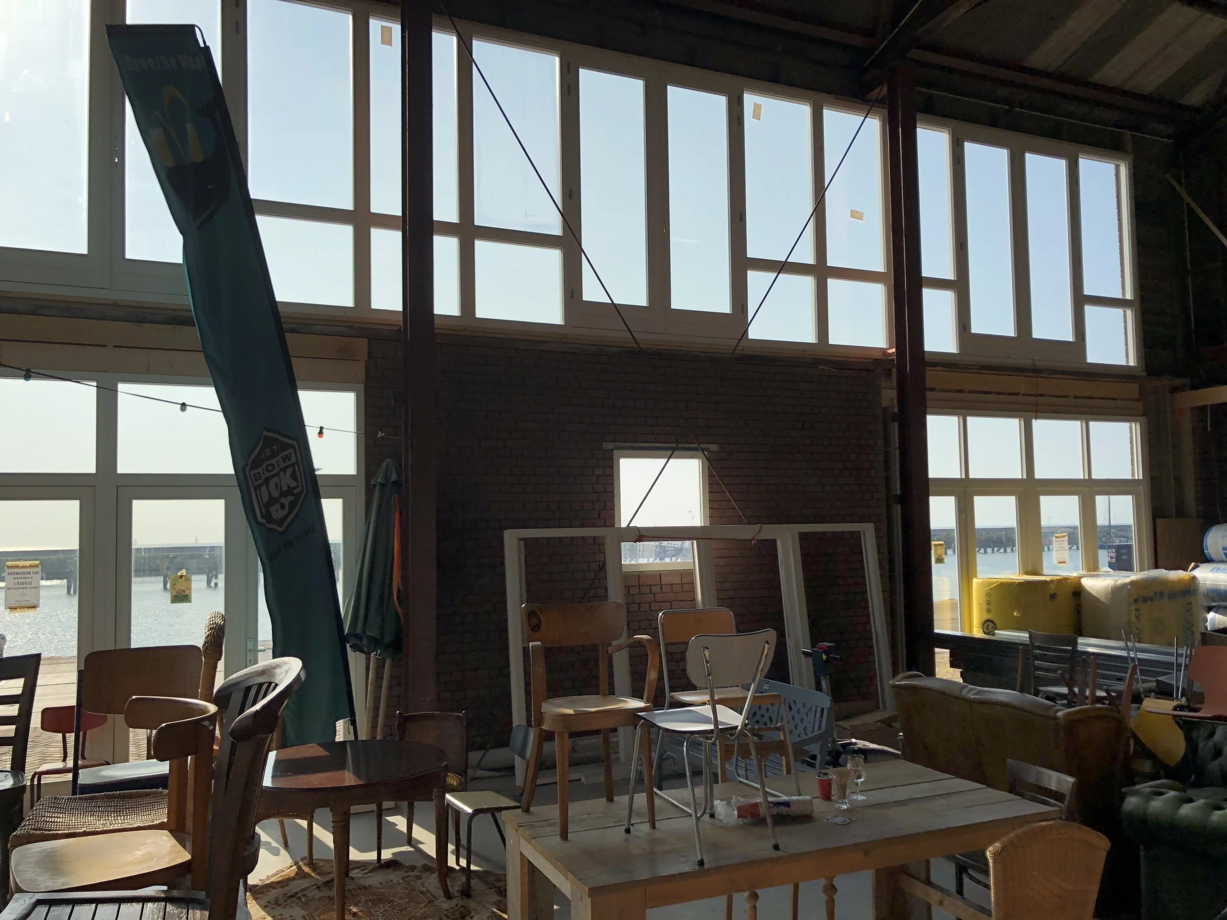 Interieur van een gebouw met grote ramen die uitkijken op een waterkant, met verschillende stoelen, tafels en bouwmaterialen binnen.