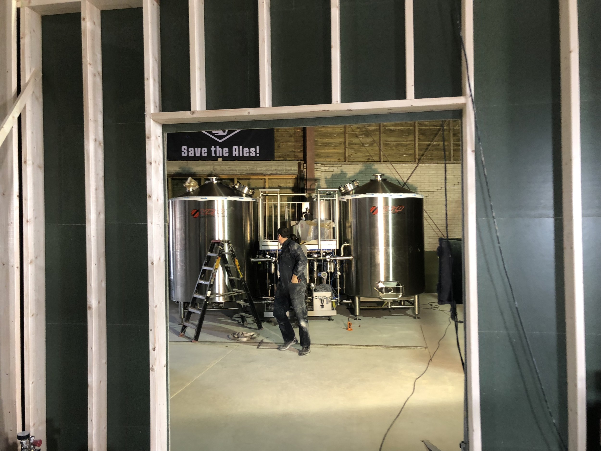 Binnen in een bouwplaats met een man die aan werkzaamheden bezig is bij grote roestvrijstalen machines, een ladder en een circuit. Er hangt een bord met 'Save the Ales!'