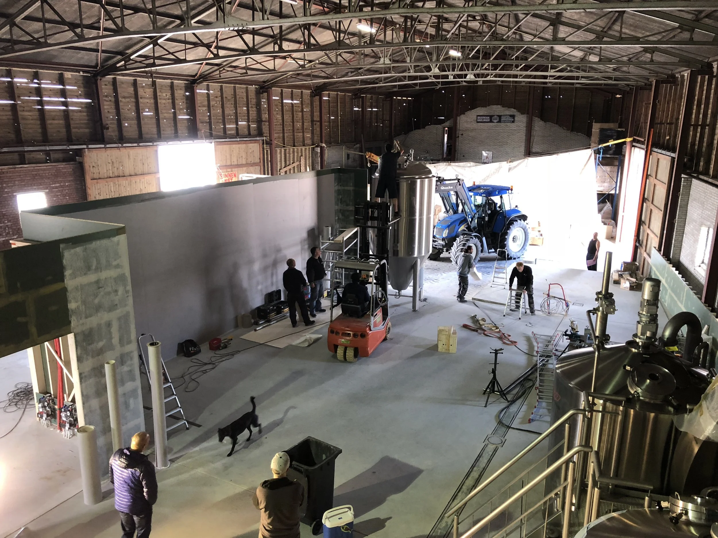 Binnen in een grote fabricagehal worden mensen en machines waargenomen, met een tractor en bouwmaterialen zichtbaar, en een persoon die op een ladder staat.