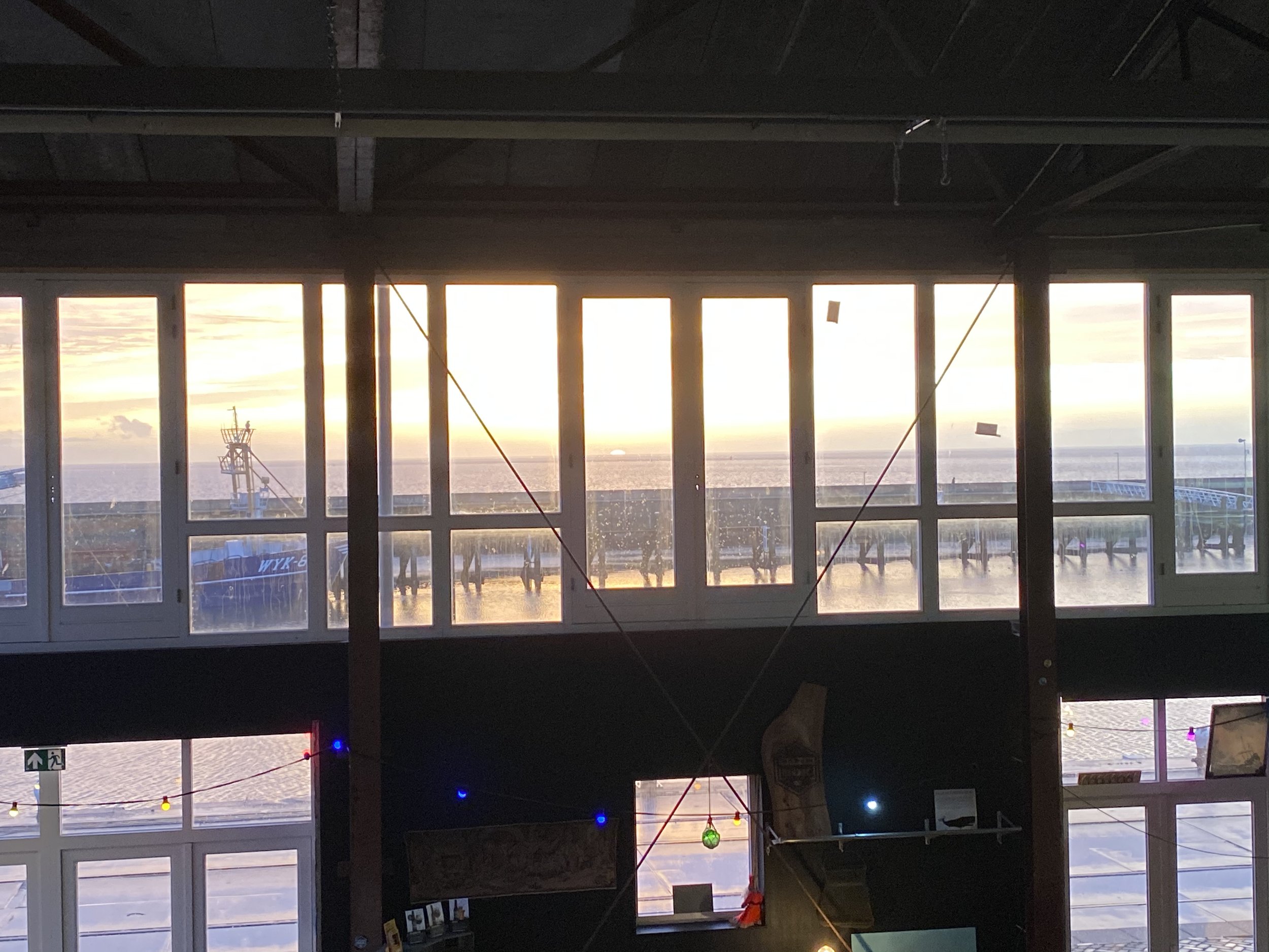 Binnenkant van een gebouw met grote ramen die uitzicht bieden op de zee bij zonsondergang, met enkele bouwmaterialen en kerstversieringen.
