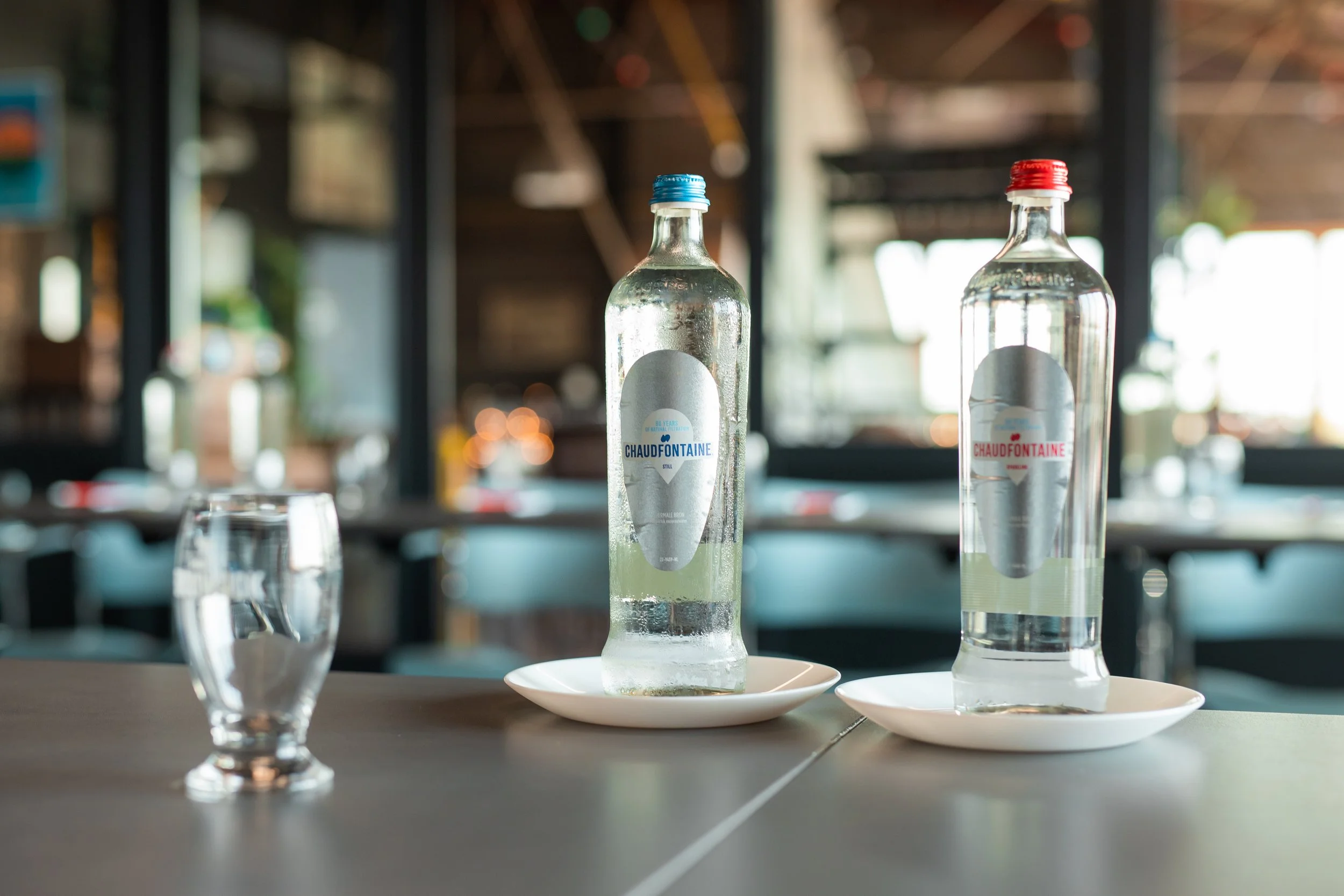 Twee heldere glazen flessen water, één met een blauwe dop en de andere met een rode dop, staan op een witte schotel op een tafel in een restaurant of café. In de achtergrond is een interieur met grote ramen en zachte verlichting te zien.