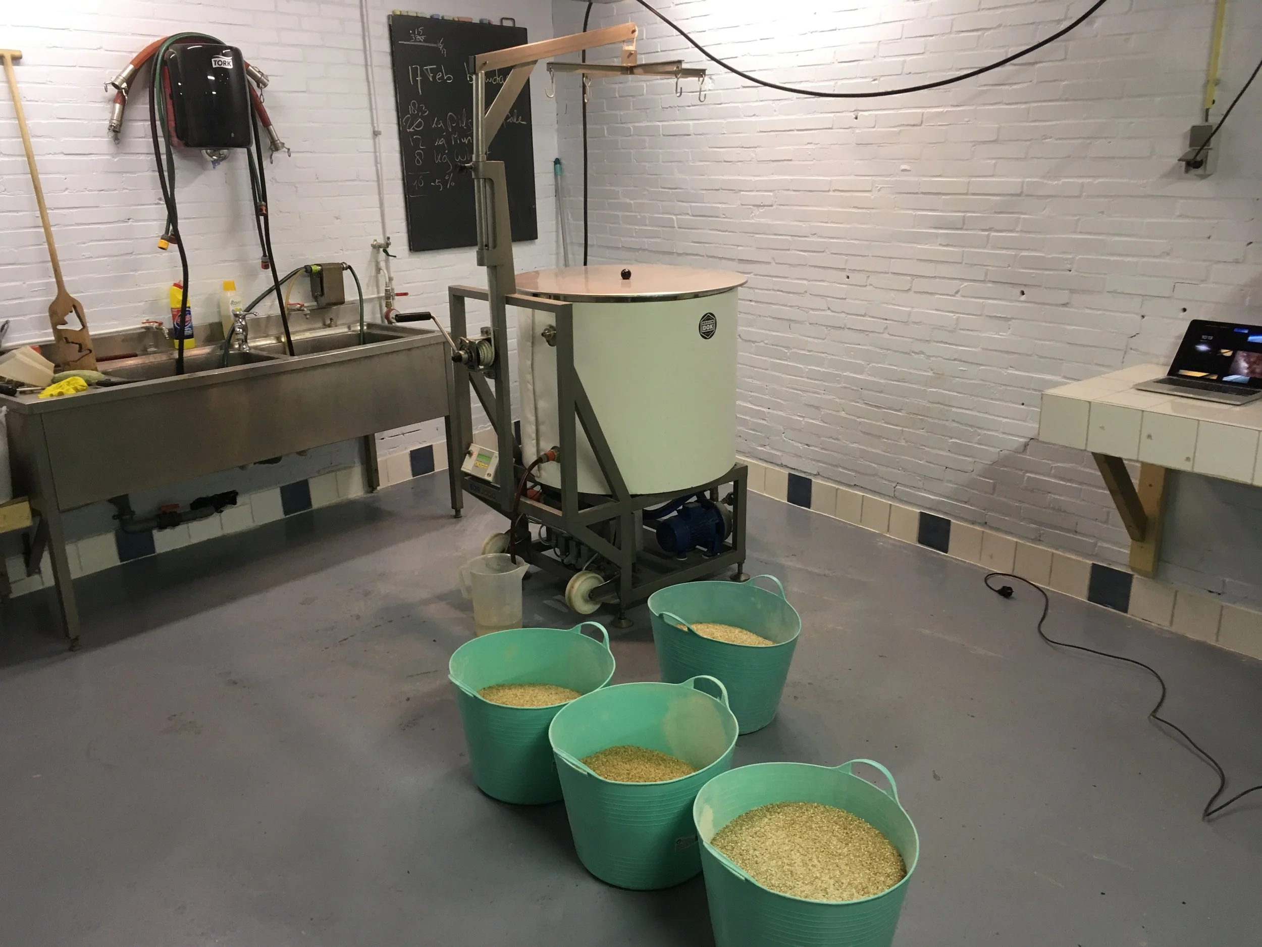 Een binnenshuisruimte met een wastafel en opslag voor schoonmaakmiddelen, een zwart krijtbord met datum en notities, een machine met een ronde deksel en een kraan, en vijf groene bakken gevuld met graan op de vloer.