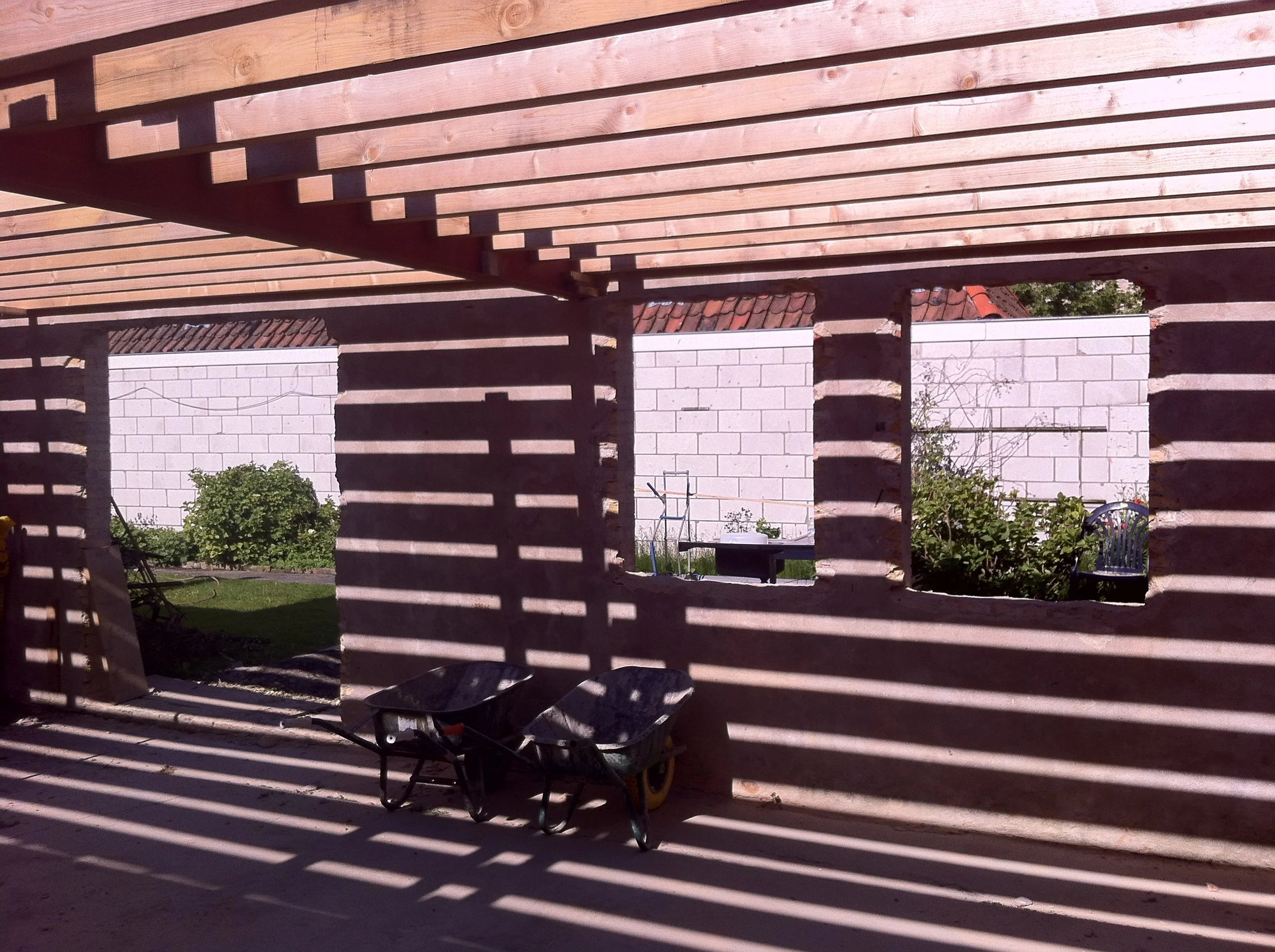 Buitenperspectief van een houten terrasconstructie met schaduwpatronen op de grond, met tuingerei en een witte bakstenen muur op de achtergrond.