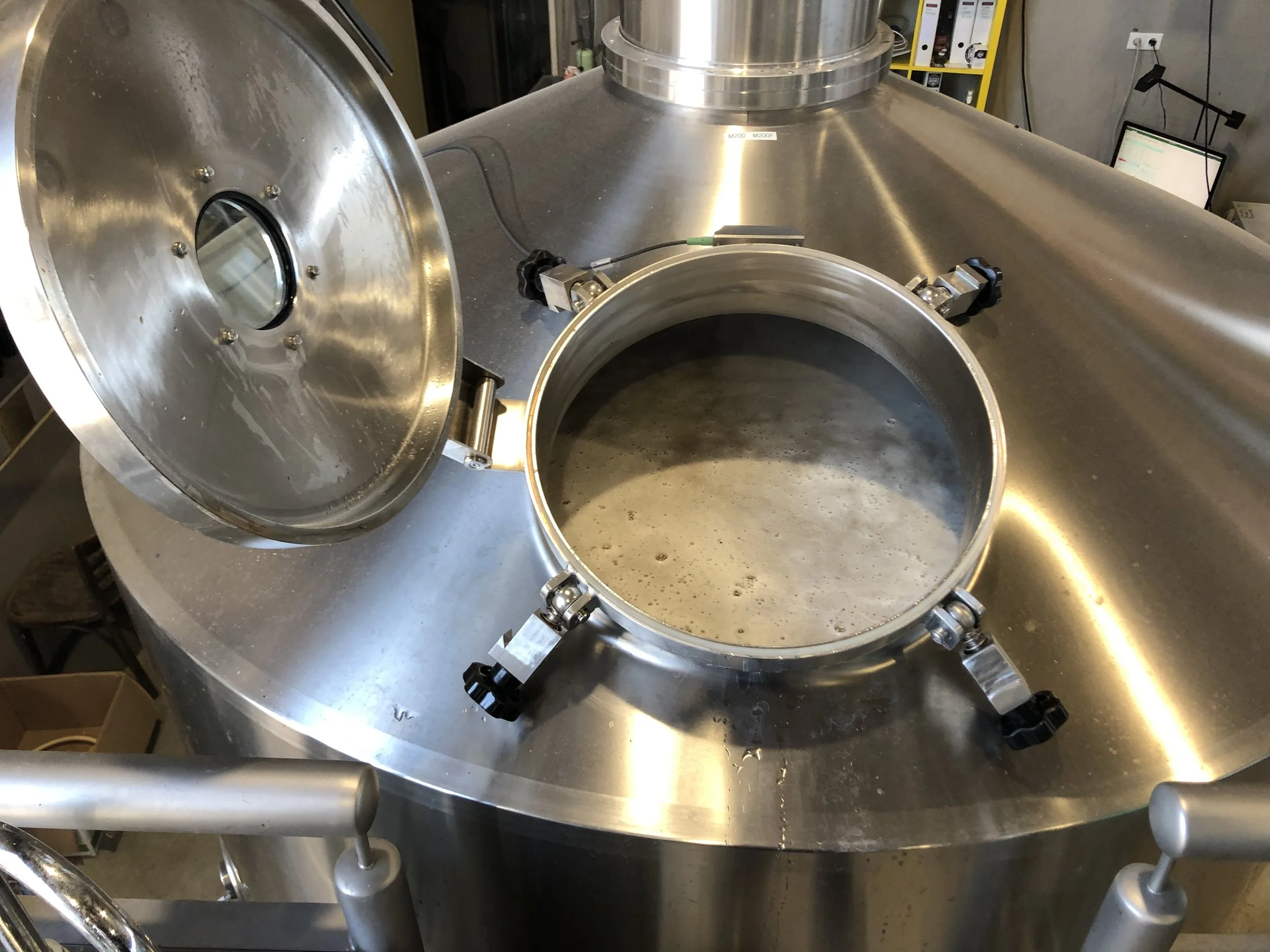 Het beeld toont een groot, roestvrijstalen instrument, mogelijk een cryogeen of een vacuümkamer, met een open deksel en een ronde opening. In de achtergrond zijn elektronische apparatuur en een scherm zichtbaar.