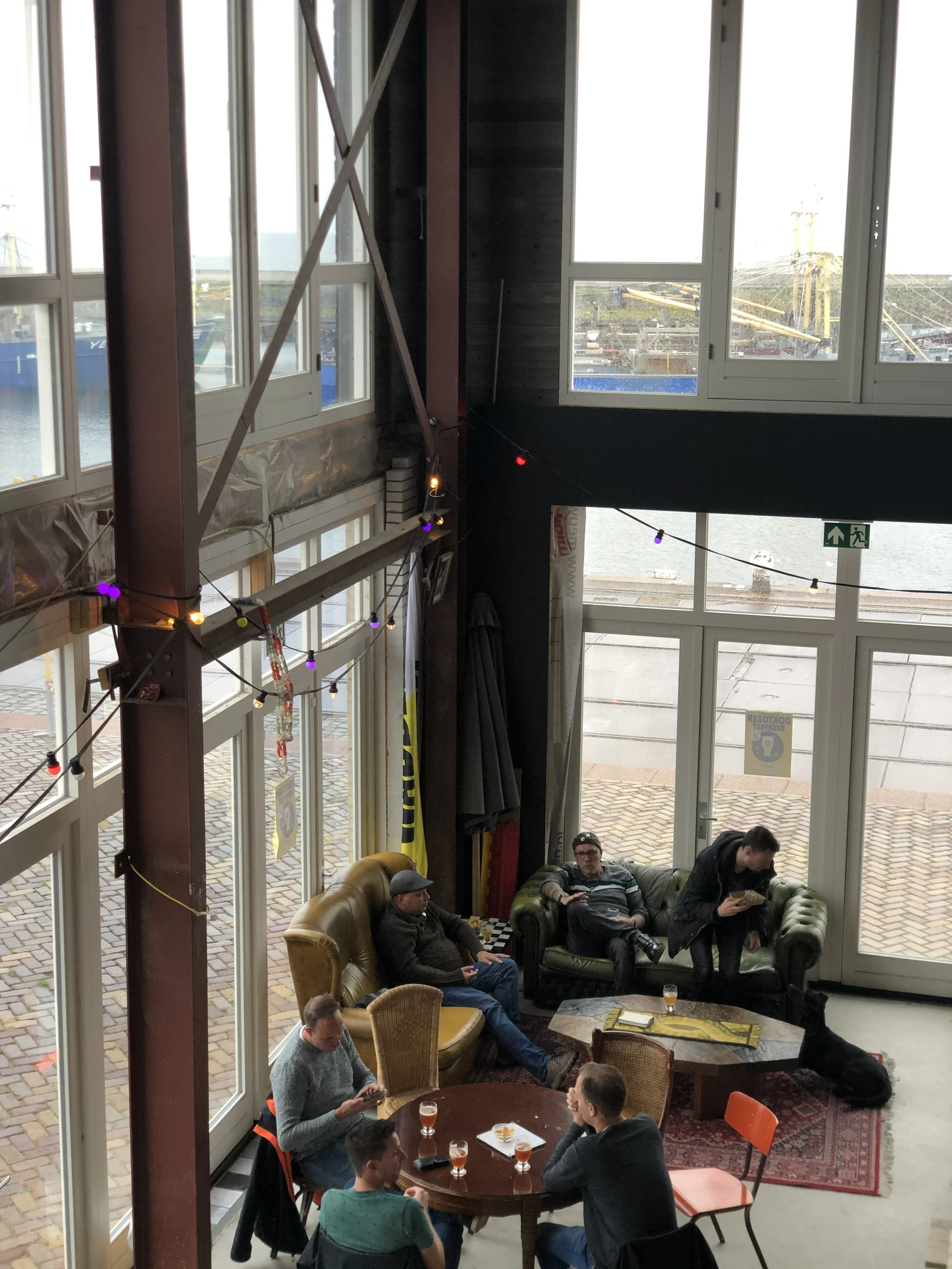 Binnenzicht van een café of woonkamer met grote ramen uitzicht op de haven, mensen zitten op banken en stoelen, sommige met drankjes, de sfeer is ontspannen.