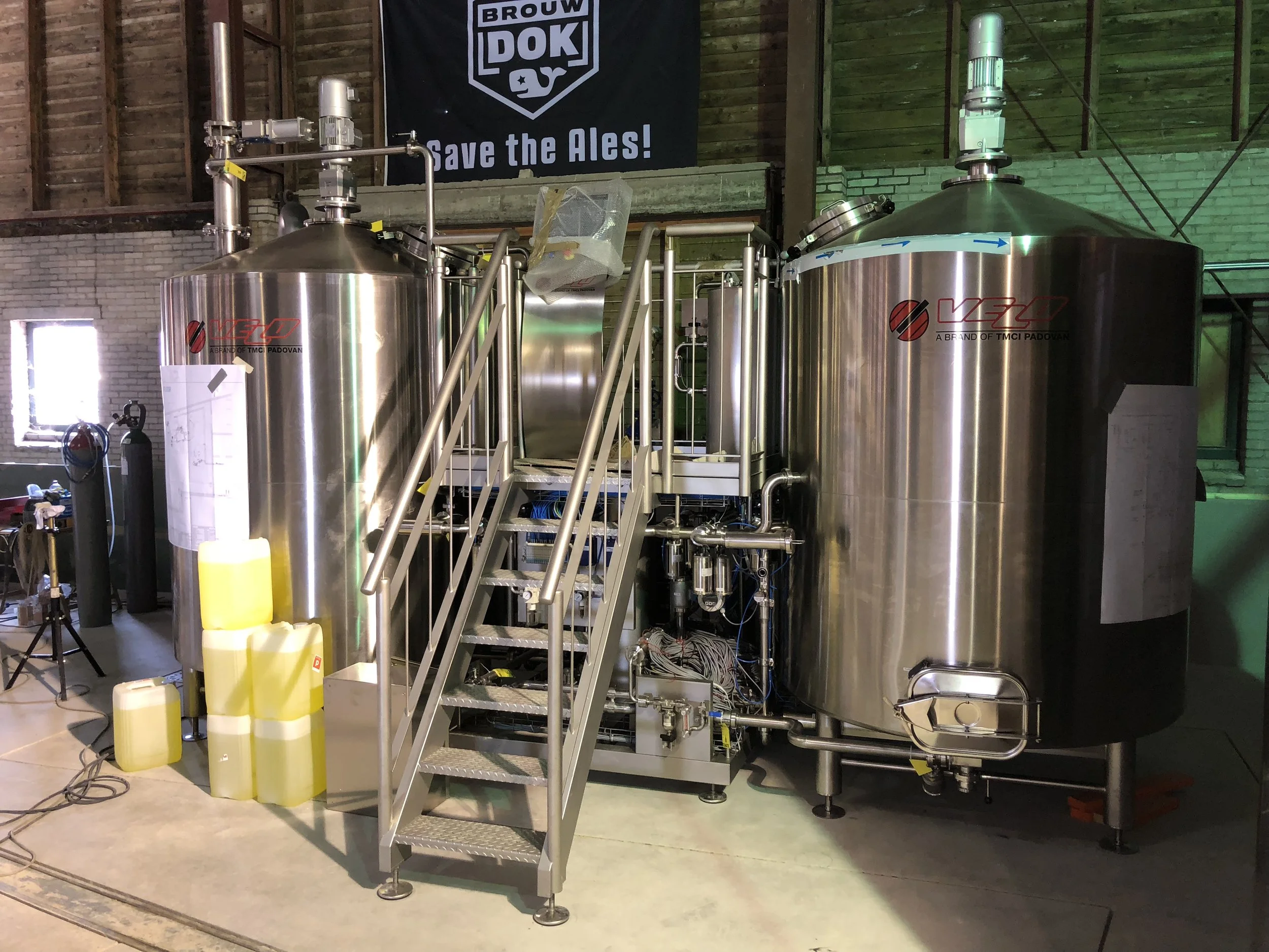 Stalen biertanks met een trap ervoor, in een brouwerij. Op de achtergrond hangt een bord met de tekst 'Save the Ales!'