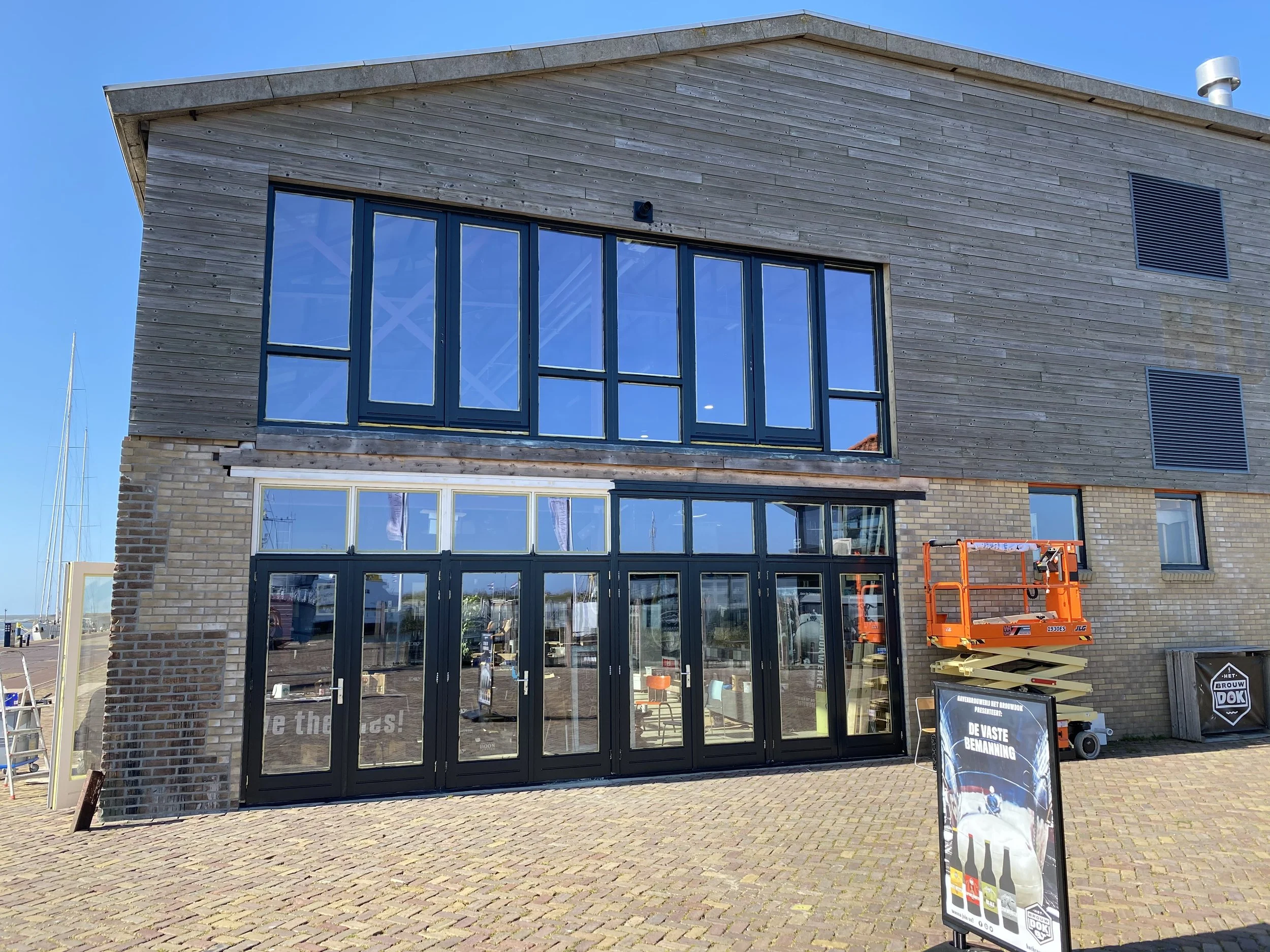 Een nieuw gebouw met grote glasdeuren en ramen, een gemengde gevel van bakstenen en hout, schilderijen en bouwgereedschap op de voorgrond, een vrachtwagen en de blauwe hemel op de achtergrond.