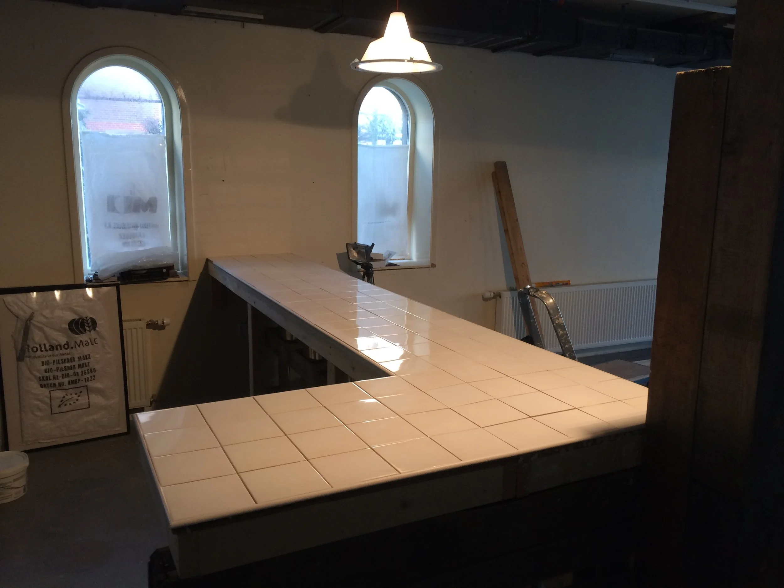 Een interieur van een kamer met twee hoge, ronde ramen, een hanglamp, een tafel met tegels en bouwmaterialen.