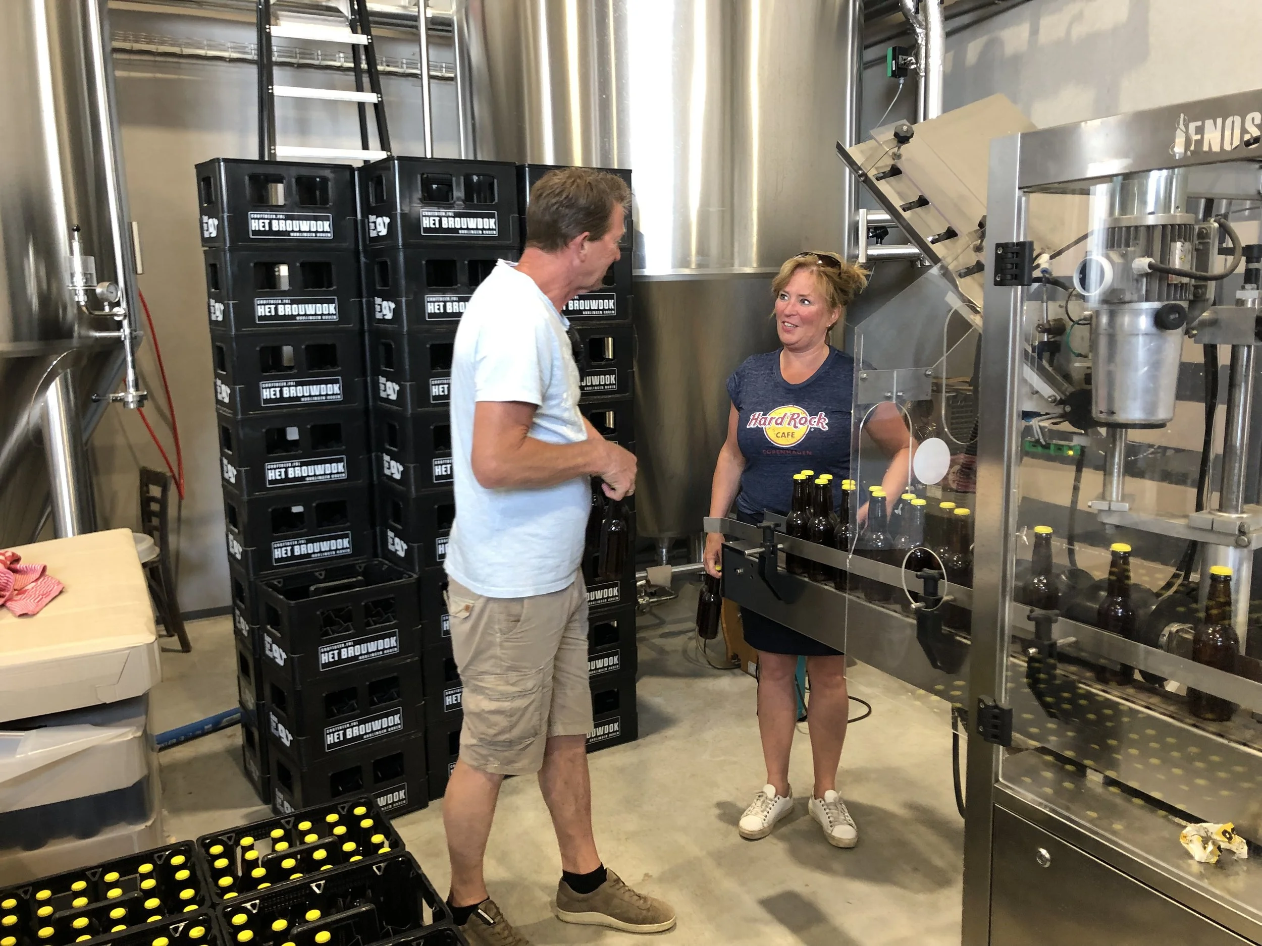 Twee mensen praten in een brouwerij. Er zijn bierflesjes, kratten en grote roestvrijstalen tanks te zien.