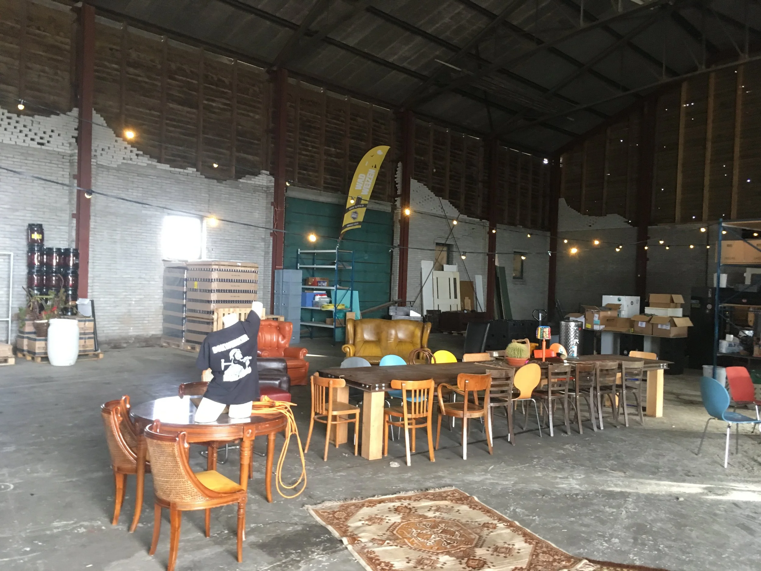 Interieur van een grote, onvoltooide of gebruikte ruimte met houten en bakstenen muren, verschillende stoelen en tafels, decoratieve lampjes en een vlag met tekst in het Engels.