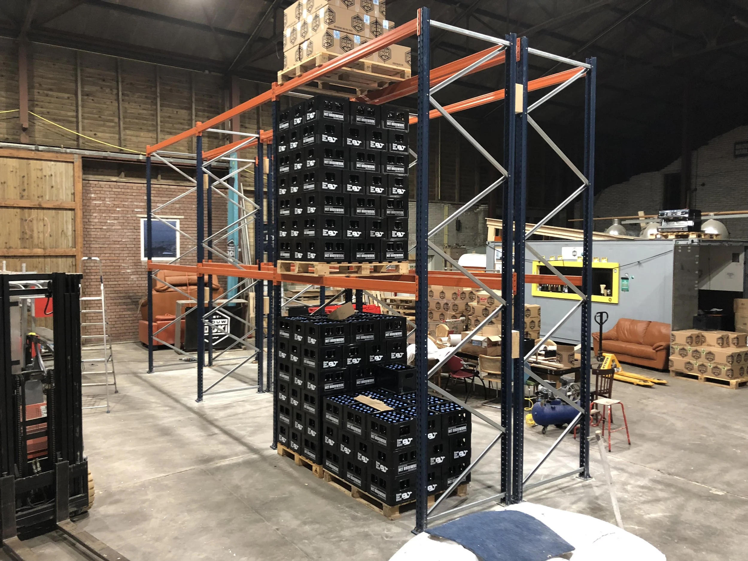 Binnen een magazijn, een metalen rek met plastic kratten en pallets met dozen, een houten bank en diverse gereedschappen en apparatuur.