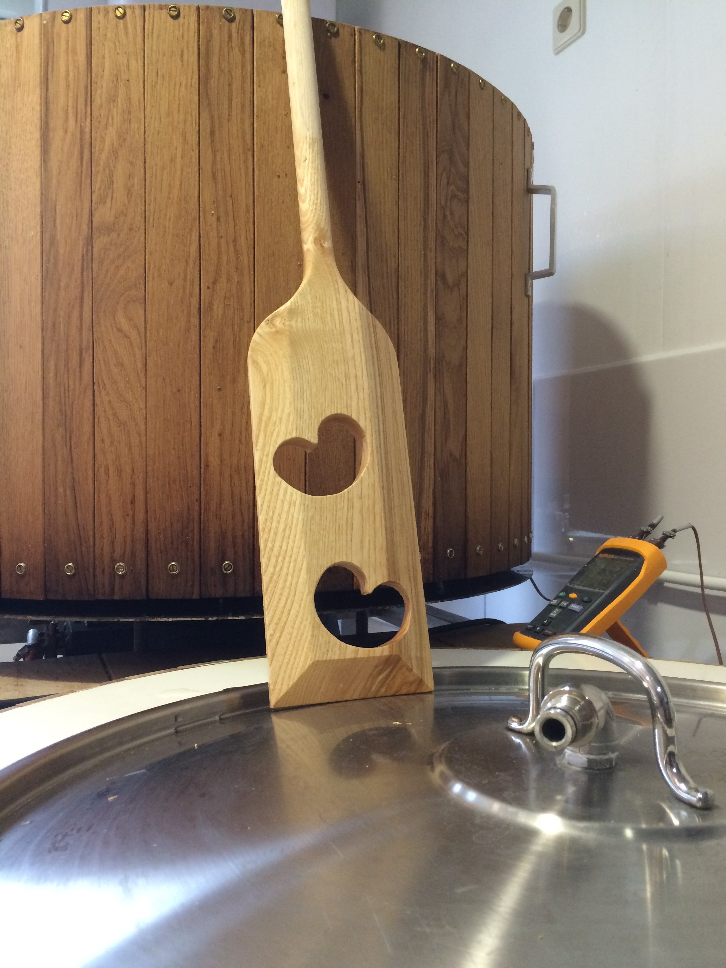 Een houten plank met een hartvormige uitsparing staat op een keuken aanrecht. Op de plank ligt een houten lepel. Op de achtergrond hangt een apparaat met draden en meetinstrumenten.