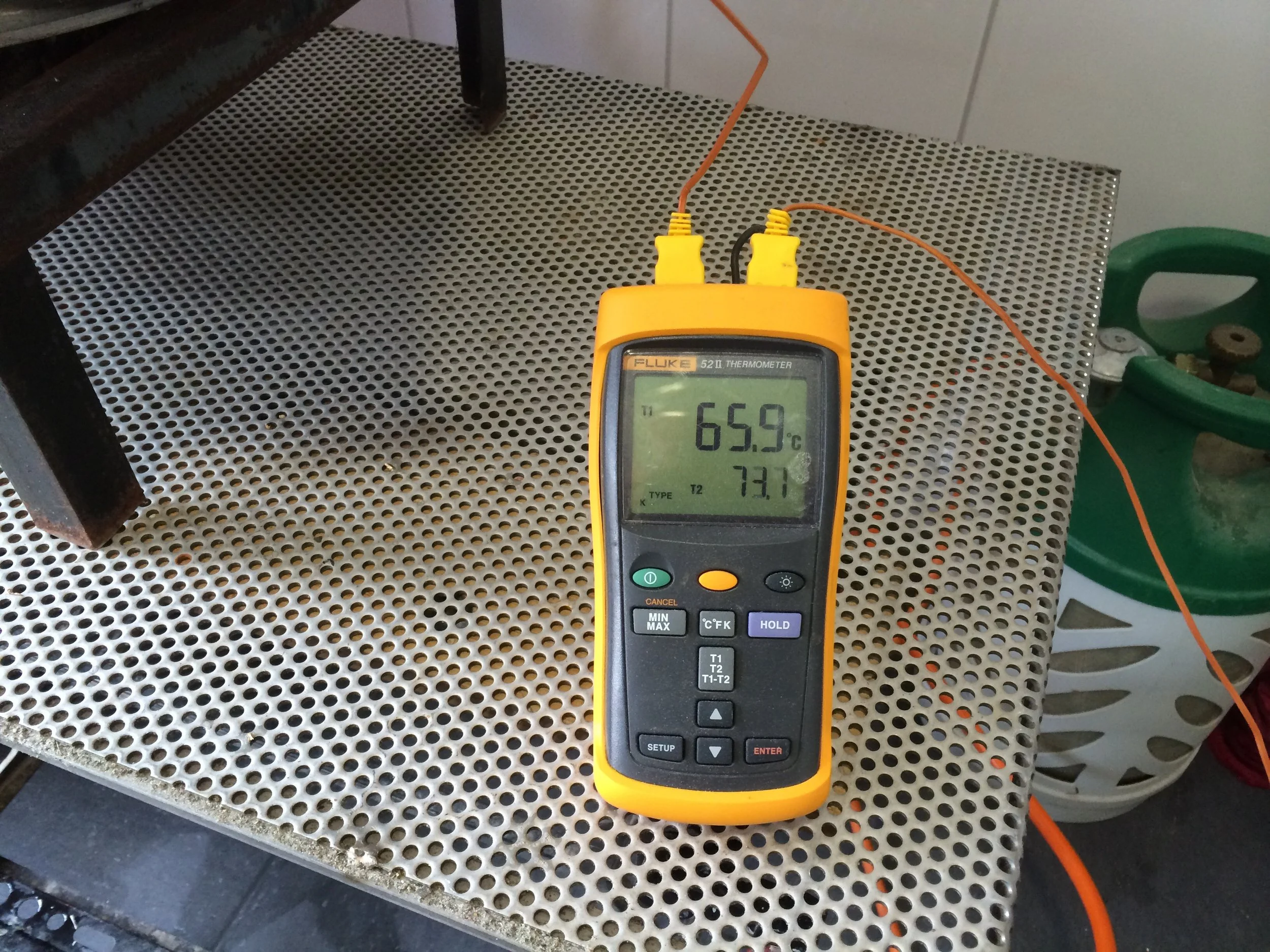 Een digitale thermometer die 65.9 graden Celsius aangeeft, ligt op een metalen geperforeerd oppervlak. Er zijn twee draden verbonden aan de thermometer, één oranje en één zwart. Op de achtergrond zijn een groene schaar en enkele kleine voorwerpen zic