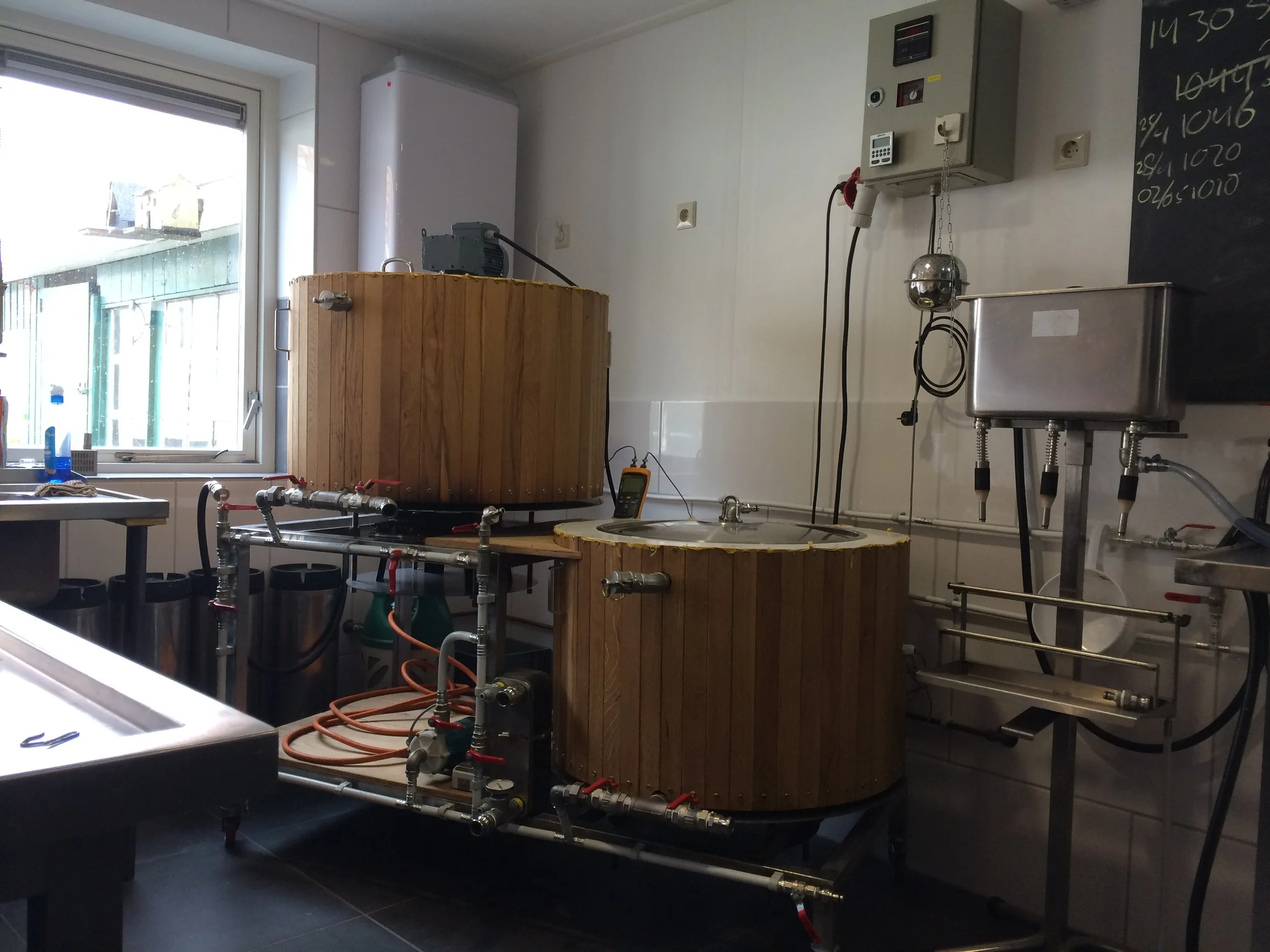 Binnen in een keuken of laboratorium met houten bakken, metalen tank, en een railsysteem van buizen en pompen, een raam en een zwart bord met witte tekst binnengezien.