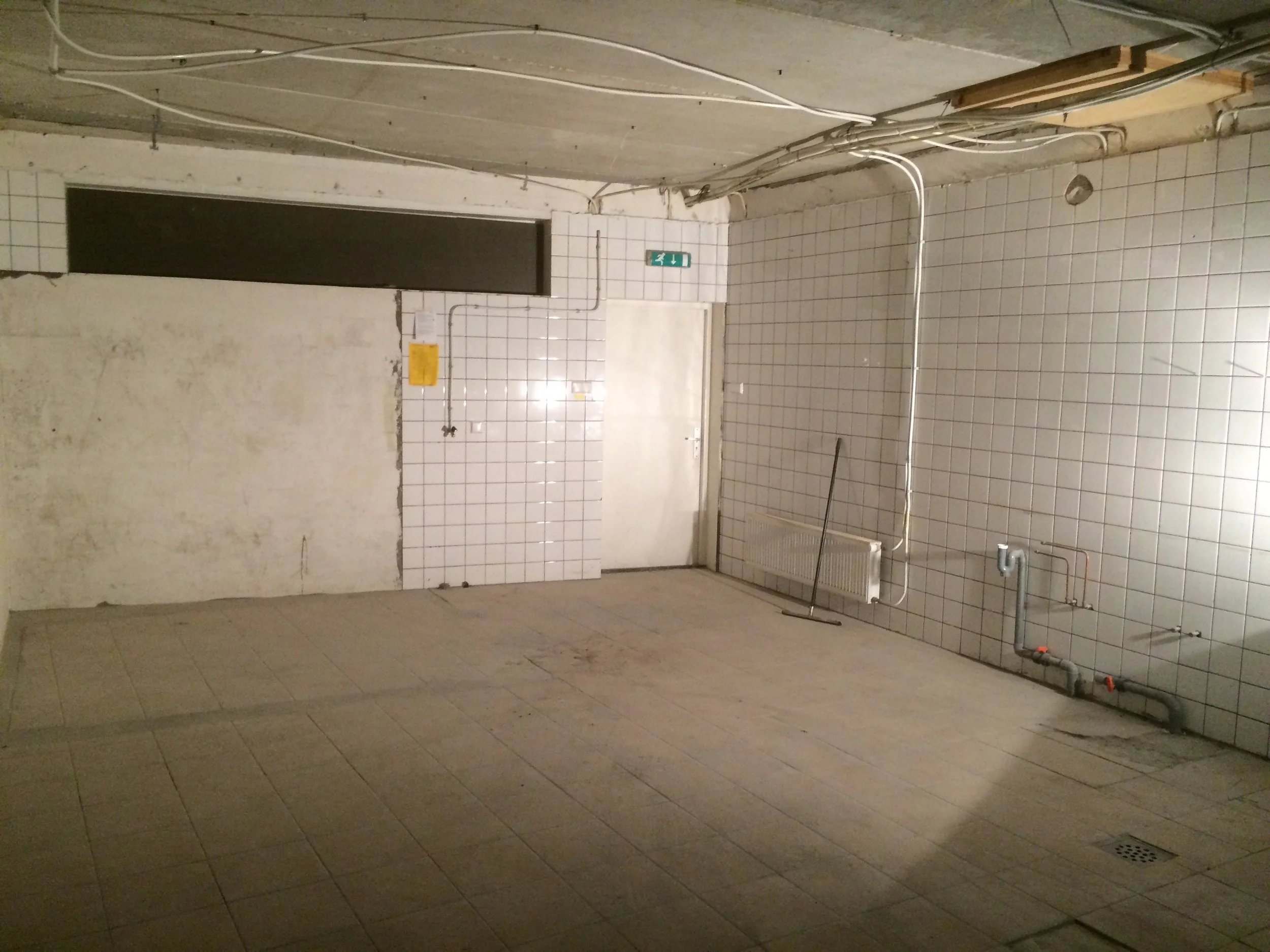 Een lege, ondergrondse toiletruimte met witte tegelwanden en een tegelvloer. Er is een nooduitgangbord en waterleidingen zichtbaar.