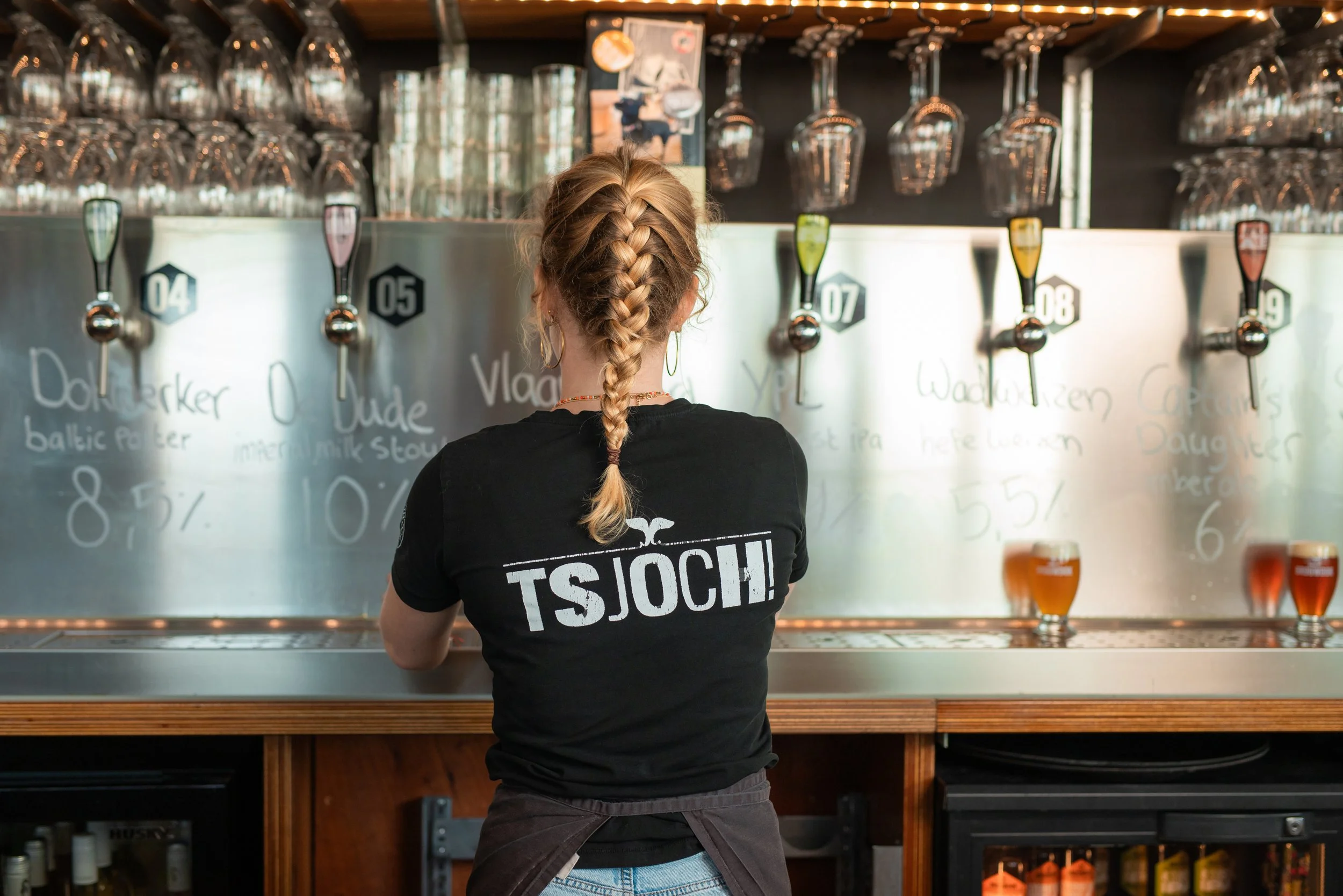 Een vrouw met een gevlochten haar draagt een zwart T-shirt en staat aan een bierkraan in een bar of brouwerij, achter haar hangen grote glazen en er staan glazen met bier op de toonbank.