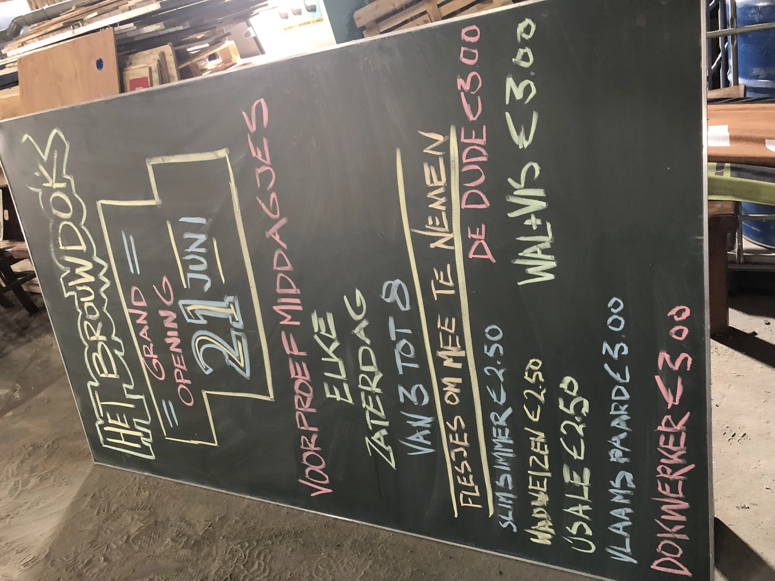 Een schoolbord met kleurrijke handgeschreven tekst die de dag van de week, de openingstijden en extra informatie over een evenement of dag aangeeft. De teksten zijn geschreven in verschillende kleuren en letters.